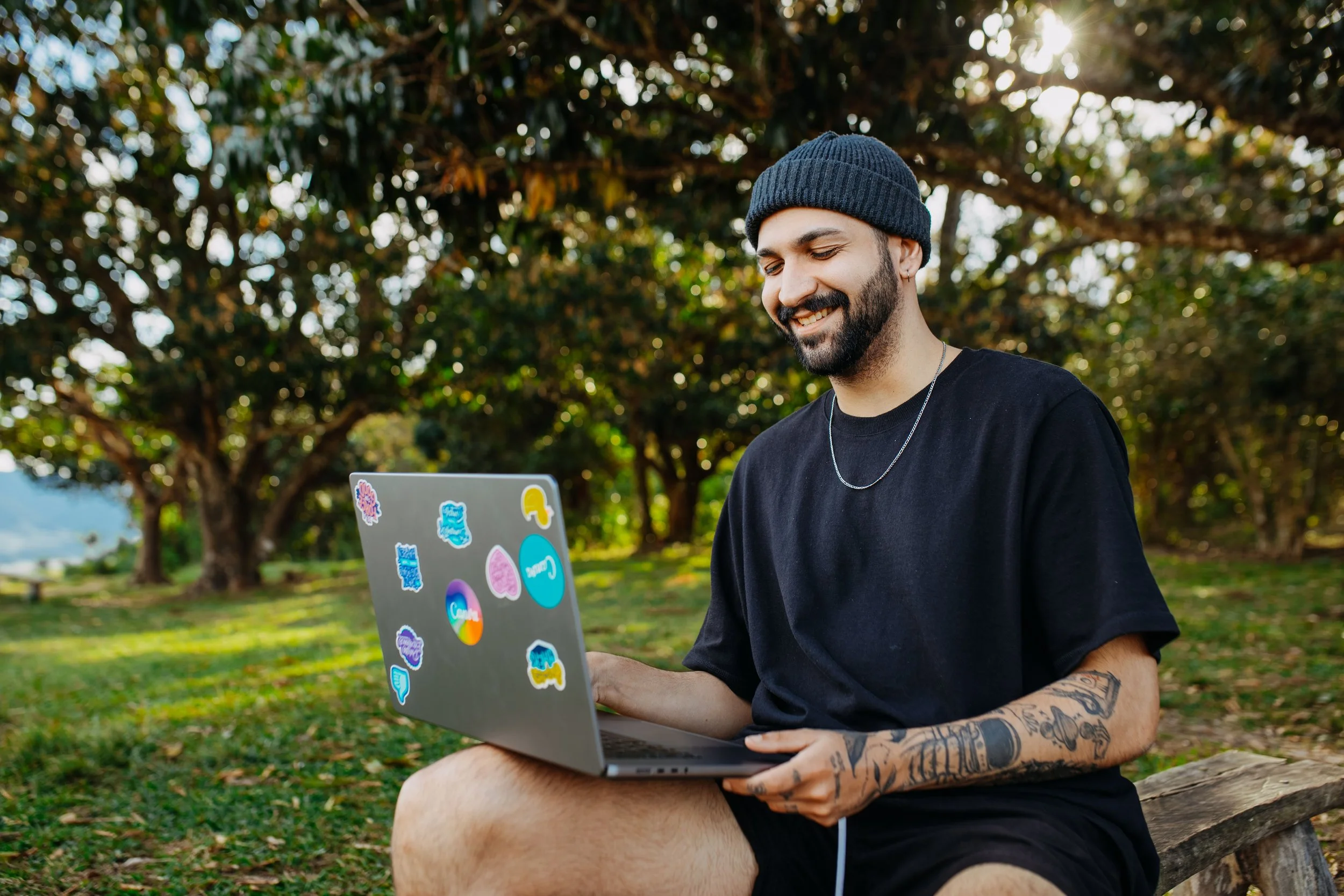 A young man with tattoos, a beard, and a beanie sitting on a park bench using a laptop decorated with colorful stickers, with trees and sunlight in the background.