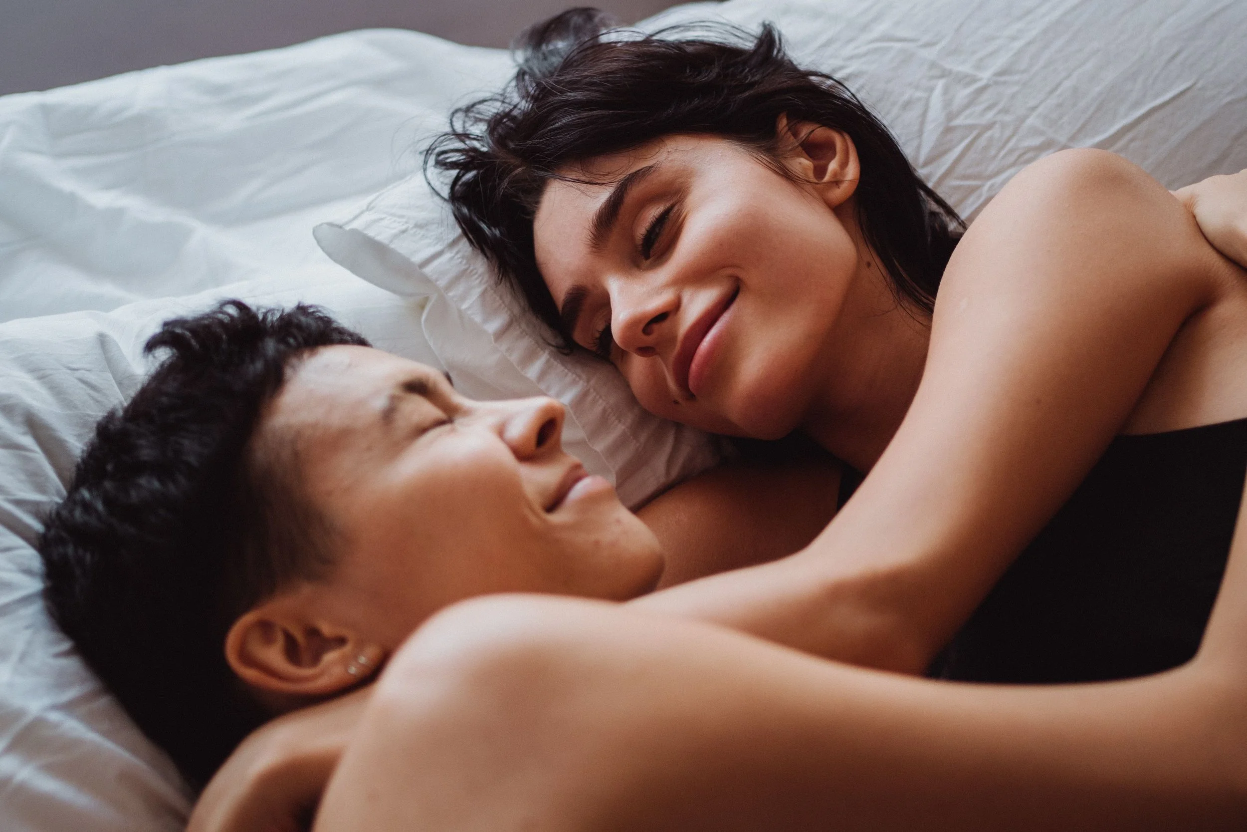 A woman smiling and cuddling with a man lying on a bed, both appearing relaxed.
