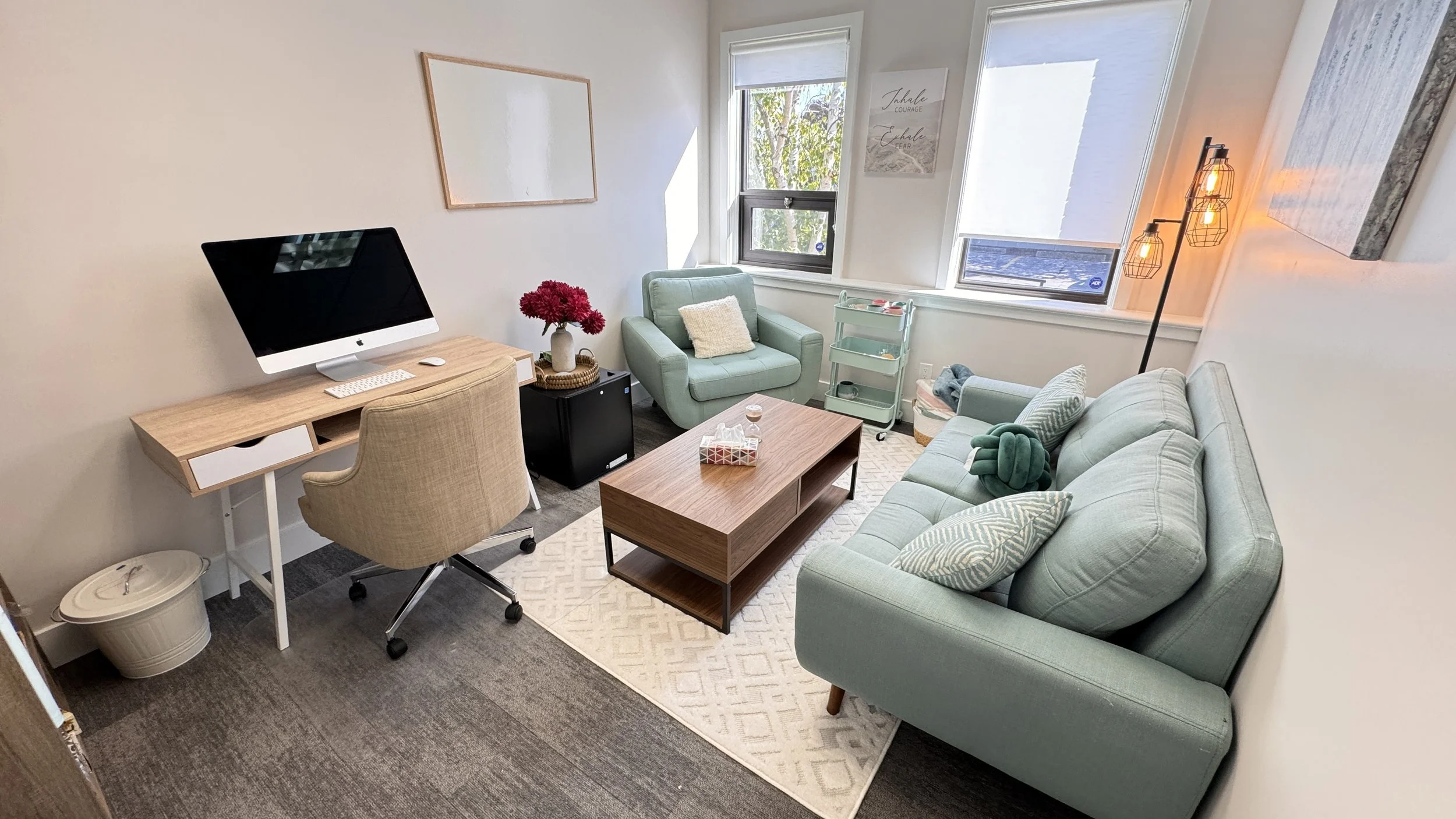 Modern home office with beige walls, featuring a wooden desk with a computer, beige swivel chair, light blue couch and armchair, wooden coffee table, floor lamp, window, and decorative items.