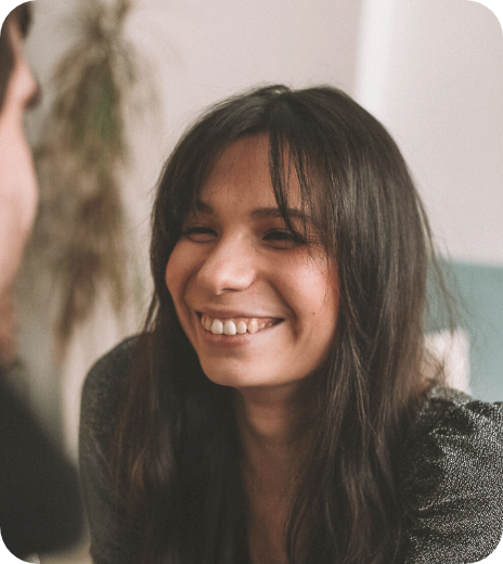 A woman with long dark hair smiling and showing her teeth, sitting indoors.