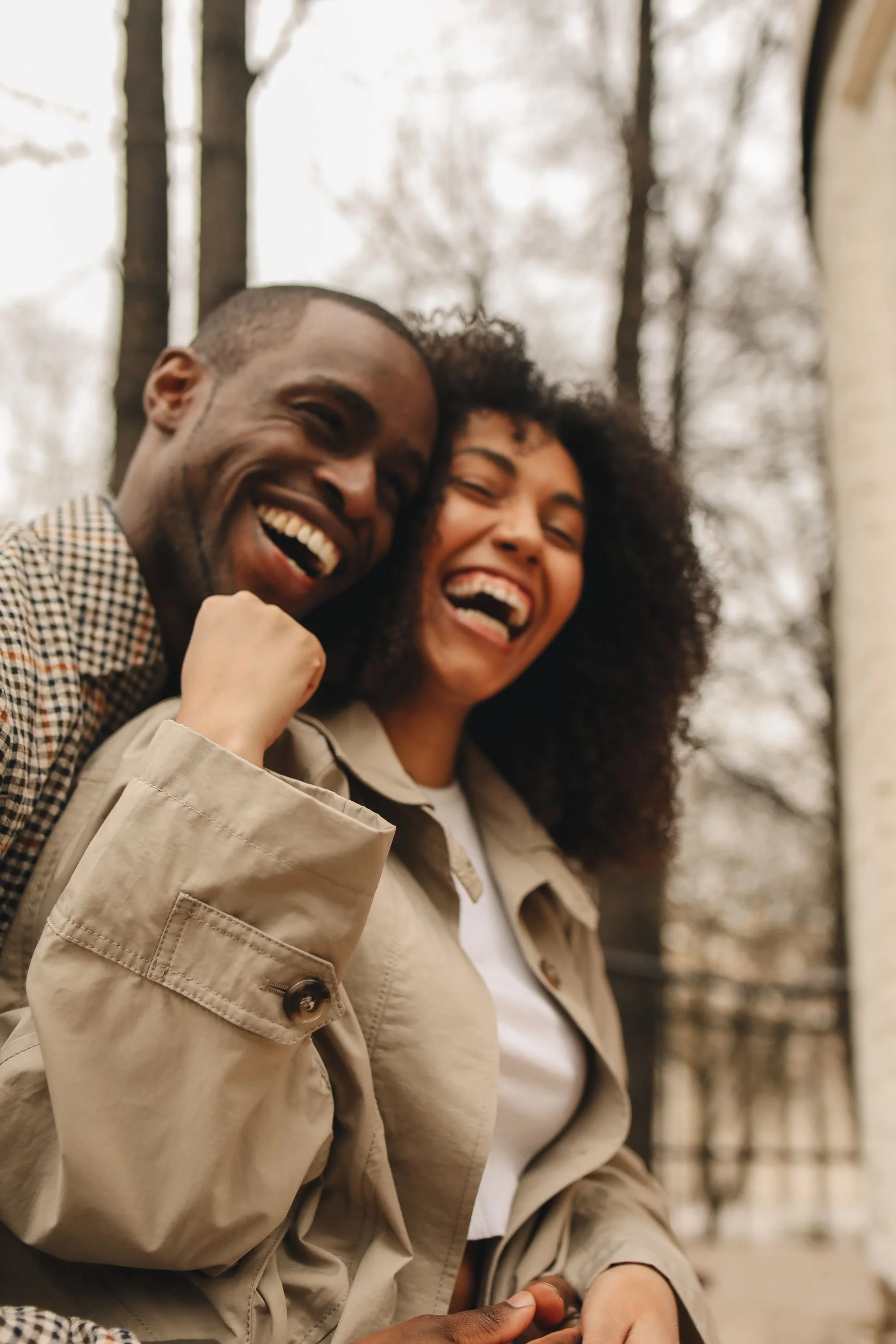 Two people smiling and laughing outdoors during daytime, with trees and a building in the blurred background.