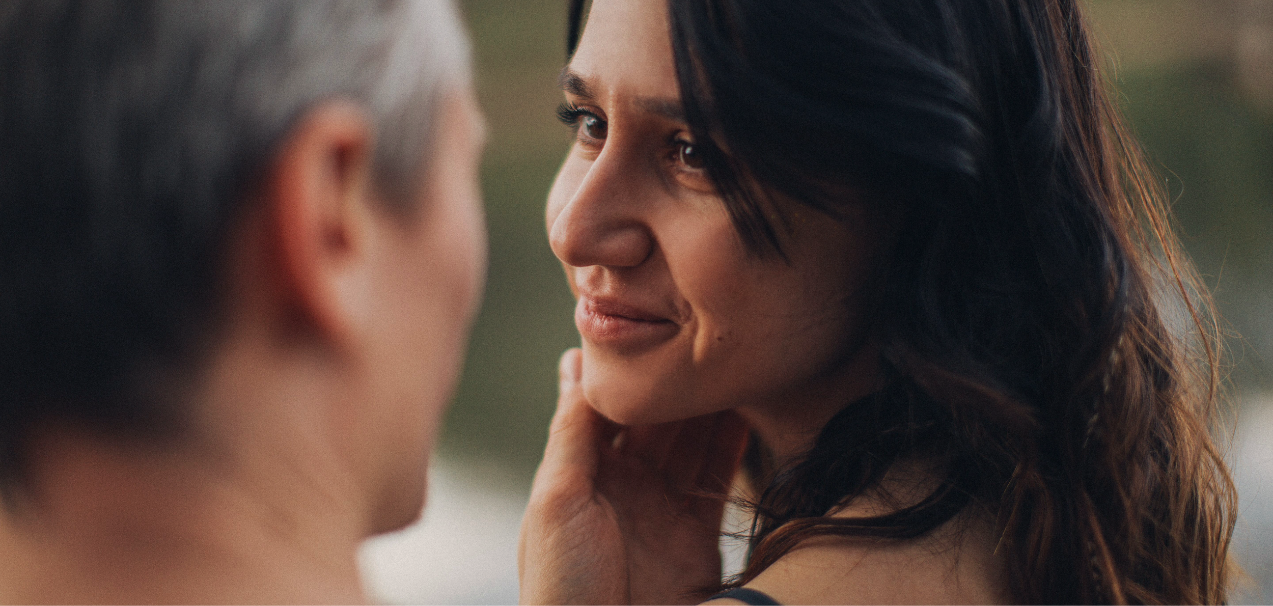 A woman with dark, wavy hair looking into the eyes of a person with short gray hair, gently touching their face.