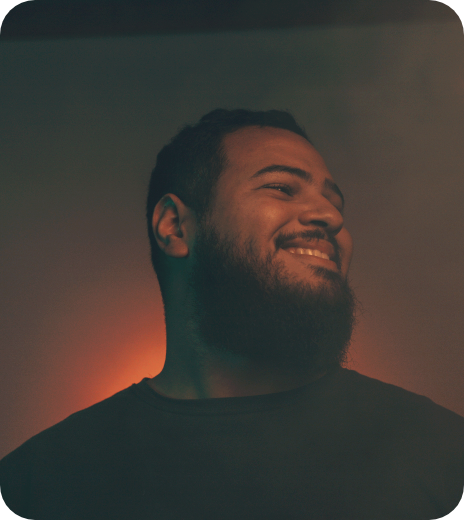 A smiling man with a beard and short hair looking to the side, dark background with warm lighting.