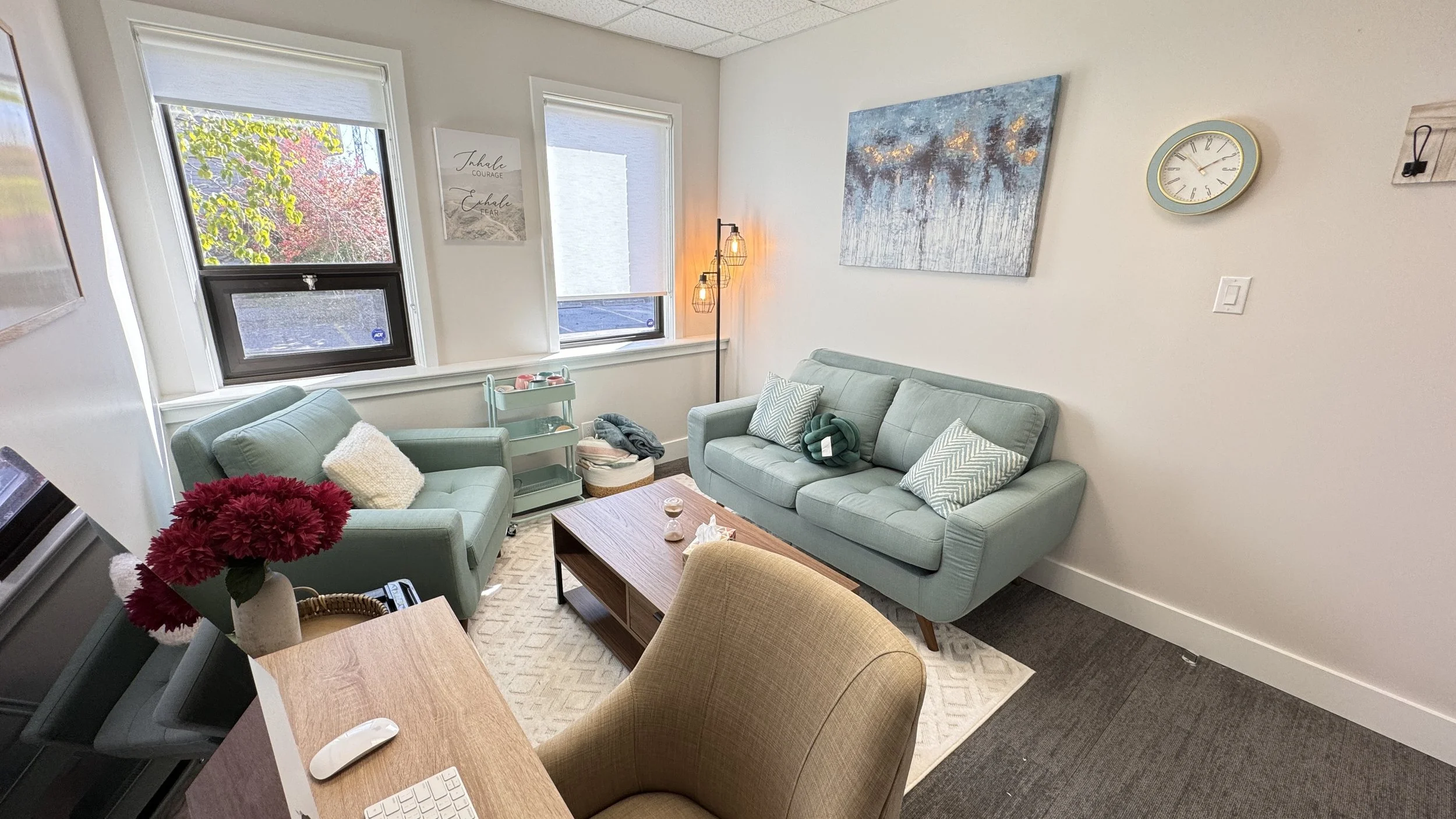 A cozy office or therapy room with light blue sofas, a wooden coffee table, a floor lamp, and wall art. A computer mouse and keyboard are on a desk in the foreground. A clock is mounted on the wall.