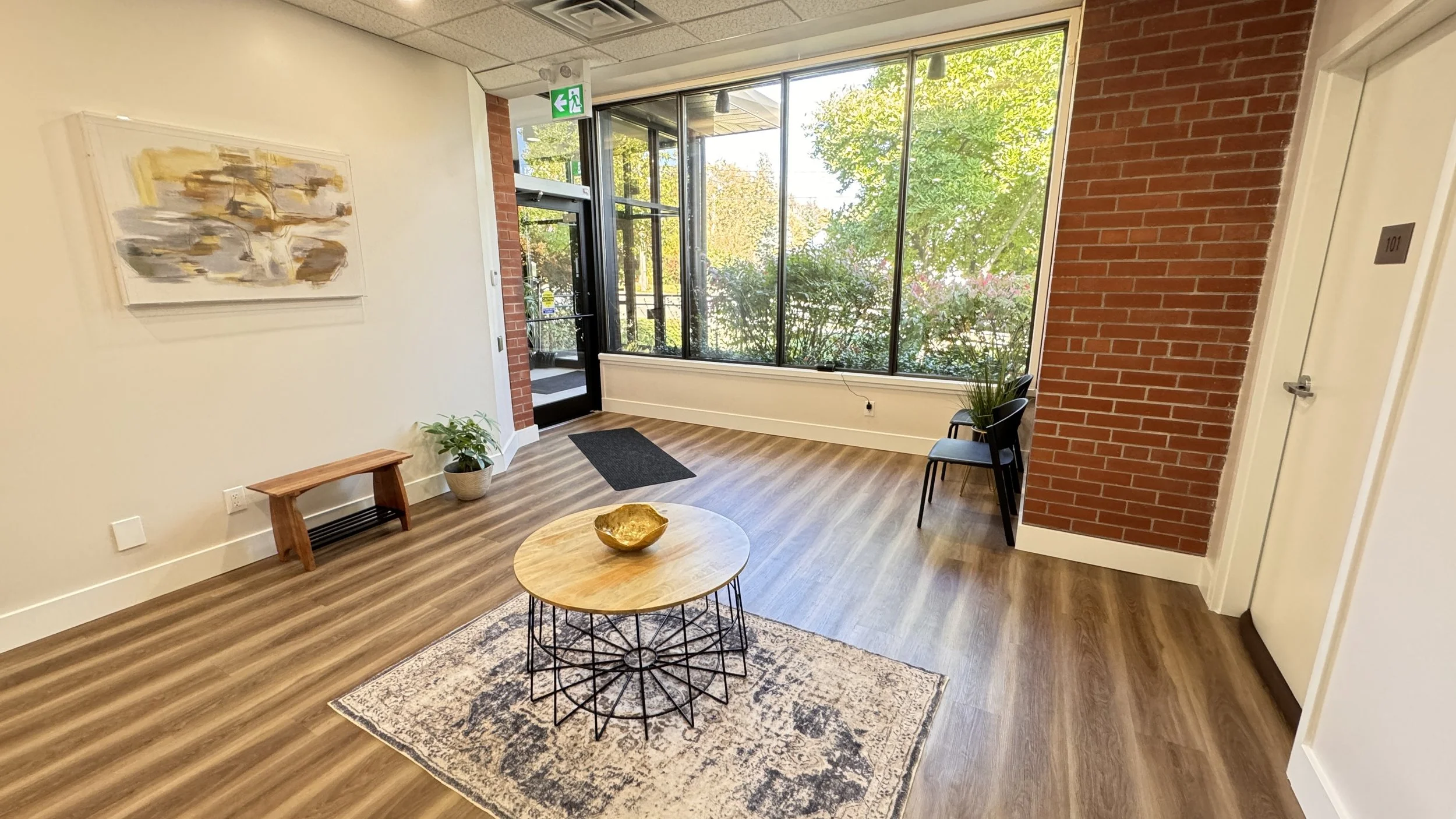 Modern office waiting area with wooden floors, circular table, chairs, plants, artwork on the wall, large windows, and brick accent wall.