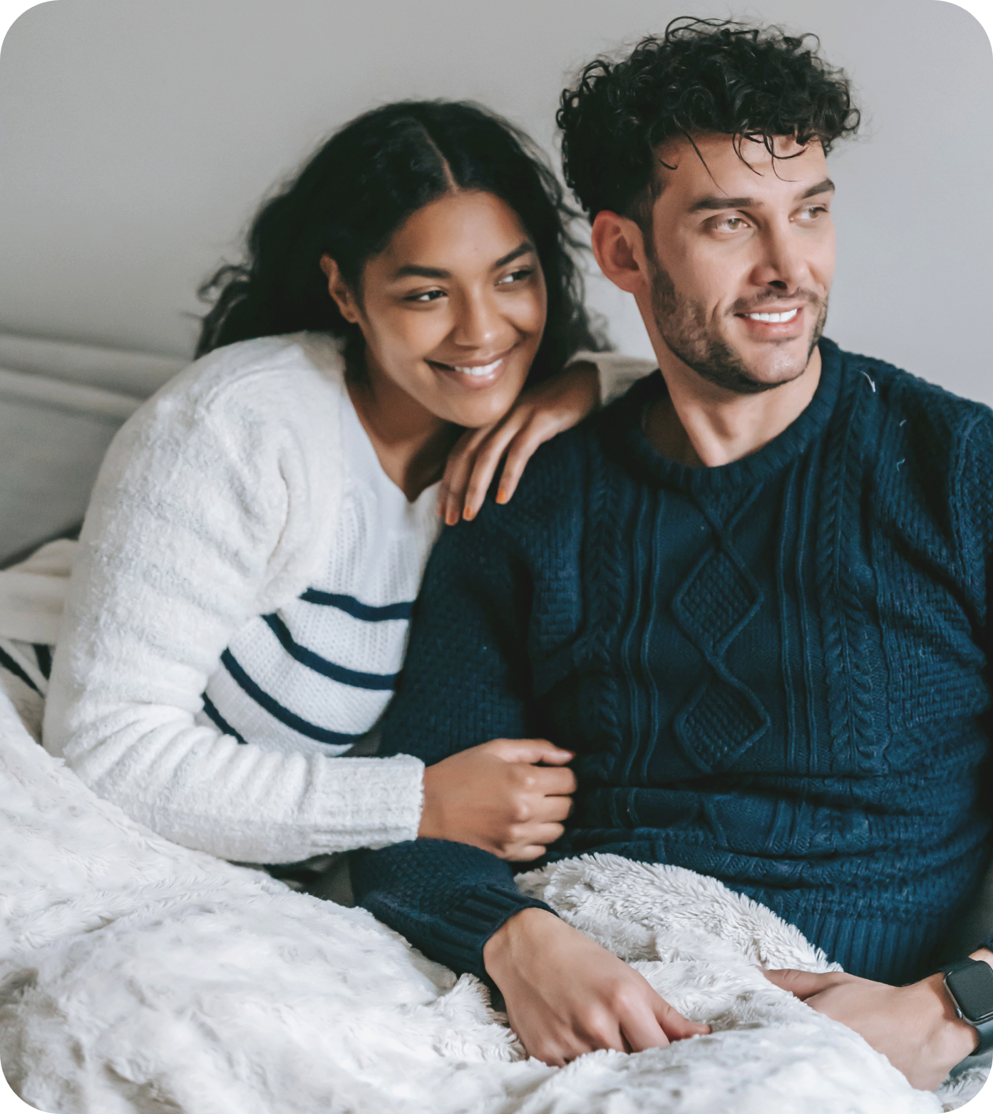 A smiling black woman with curly hair leaning on a smiling white man with curly hair, sitting on a bed with a white furry blanket, in a cozy room.