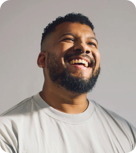 A smiling man with a beard and short curly hair wearing a light gray t-shirt, looking up against a plain background.