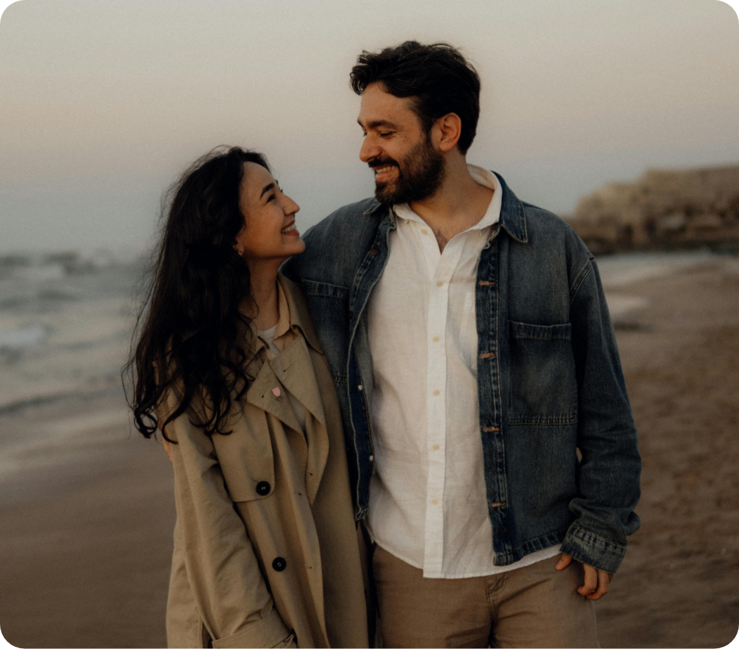A smiling couple walking on the beach at sunset, facing each other, with the ocean and rocky cliffs in the background.
