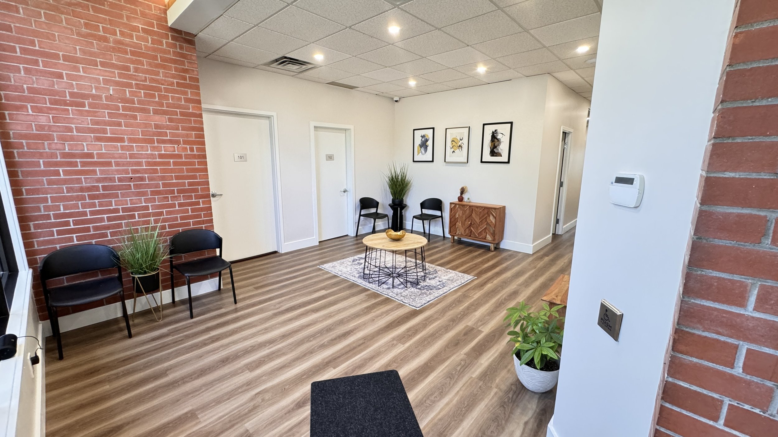 Modern waiting room with brick accent wall, black chairs, wooden table, decorative plants, artwork on walls, and wood flooring.
