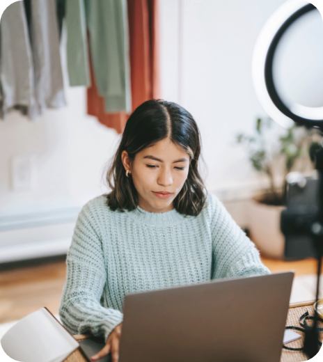 Young woman with shoulder-length dark hair using a laptop in a room with clothing hanging in the background and a ring light setup for recording or streaming.