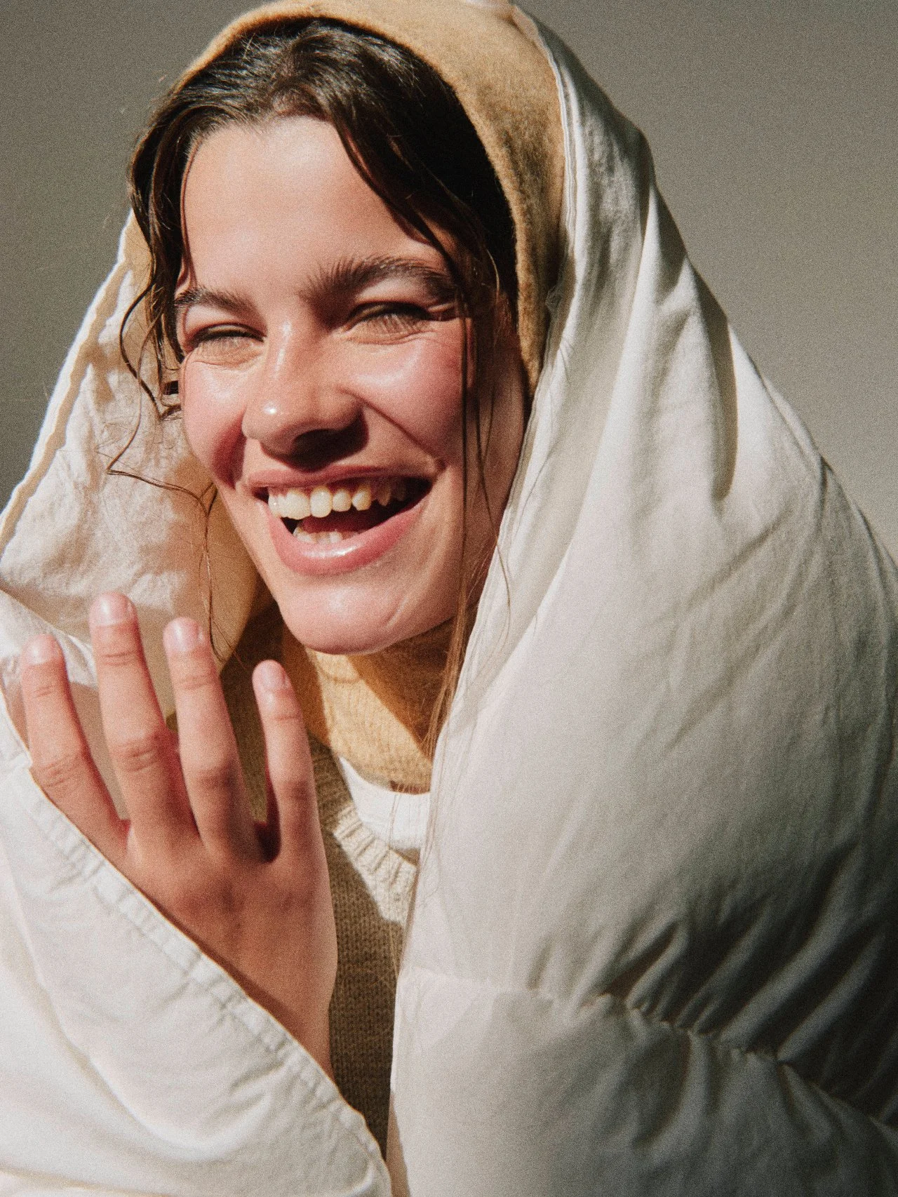 A woman with light skin and dark hair, smiling and laughing, wearing a beige hoodie with the hood pulled over her head, partially covering her face and hands.