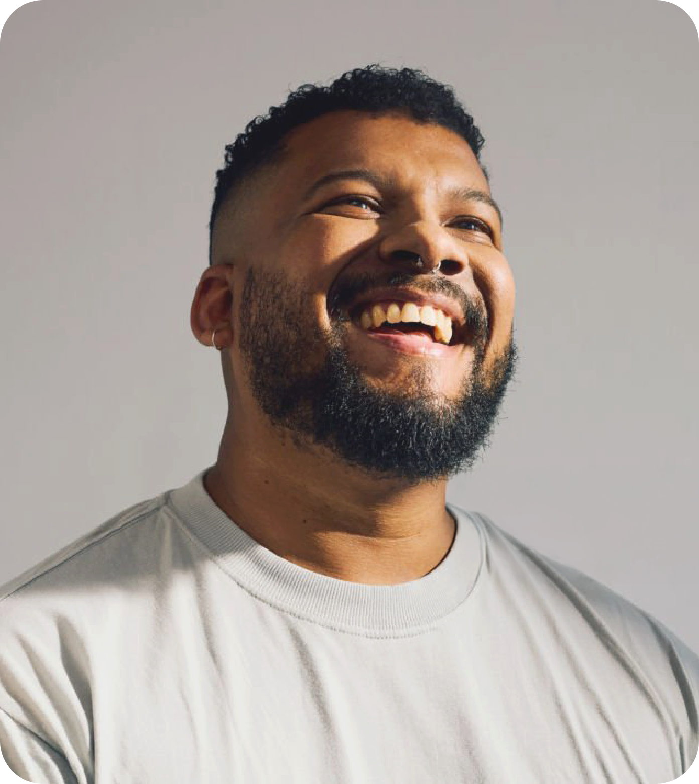 A smiling man with a beard and short curly hair, wearing a light gray t-shirt, standing against a plain gray background.