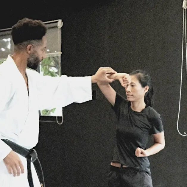 A martial arts instructor demonstrating a technique to a woman in a black t-shirt in a dojo with black walls.