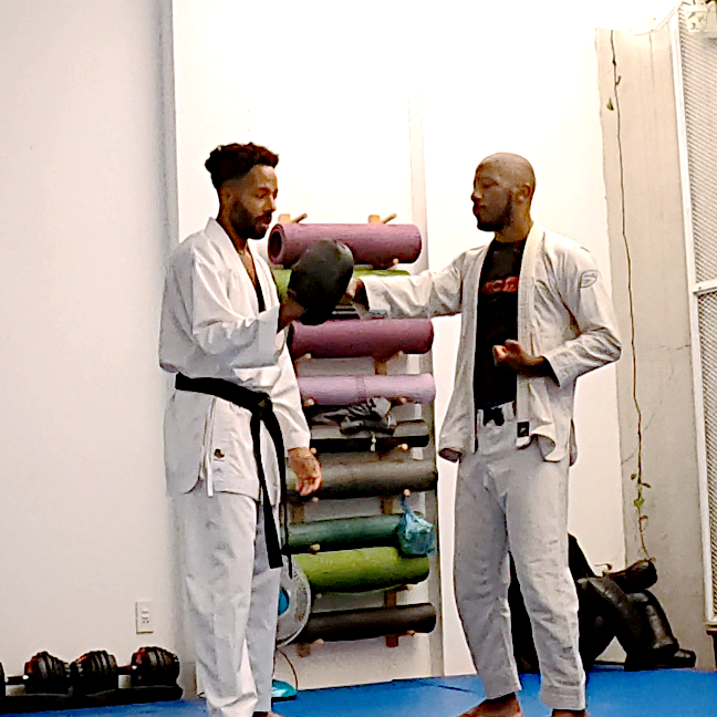 Two men dressed in martial arts uniforms practicing in a gym, one holding a boxing mitt and the other in a defensive stance, with gym equipment like mats and weights visible in the background.