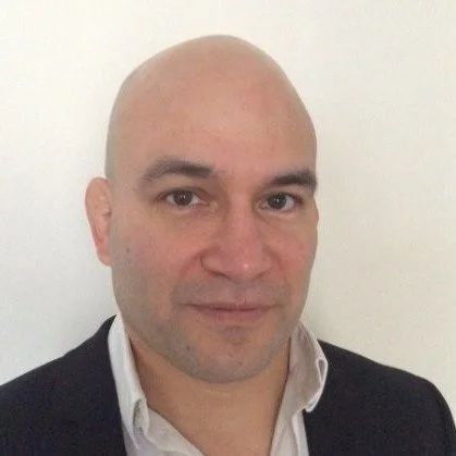 A bald man wearing a white collared shirt and a dark blazer posing for a photo against a plain white wall.