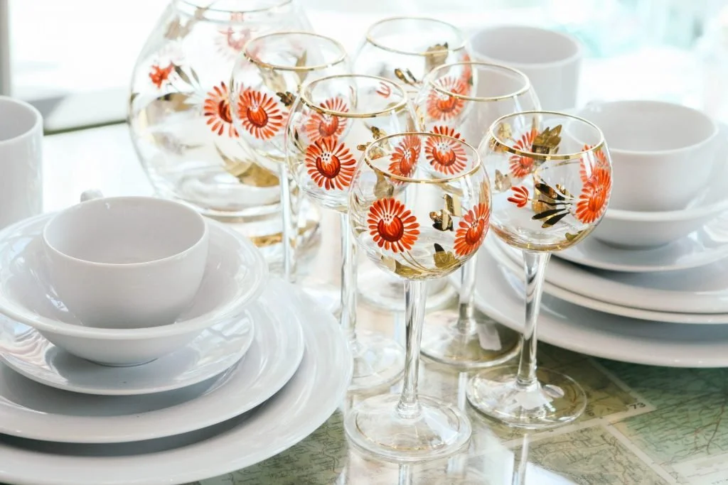 6 wine glasses with painted flowers on them next to a stack of white dishes.