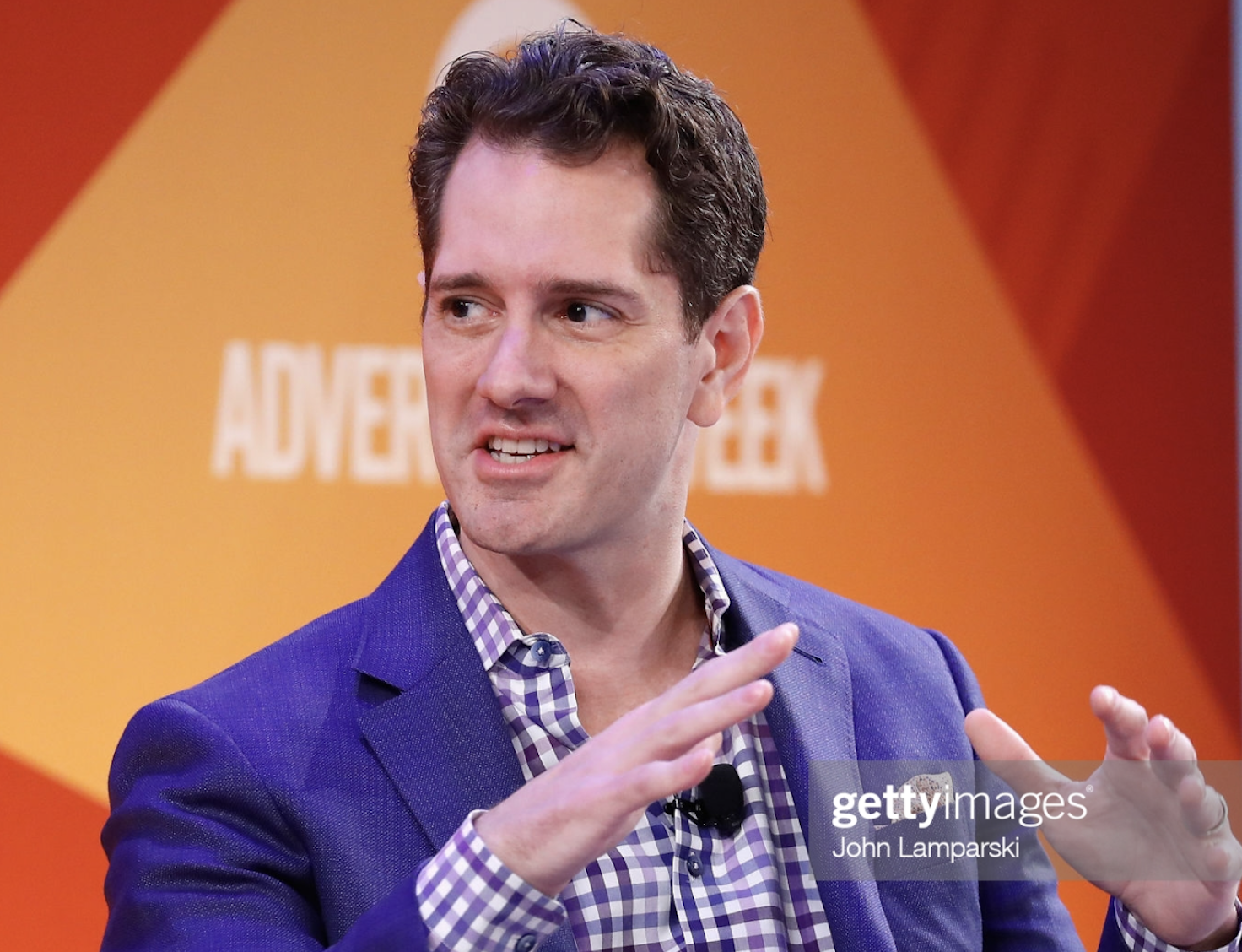 Man in blue blazer and checkered shirt speaking during an event with a yellow and orange background that has the word 'ADVENTURE' on it.