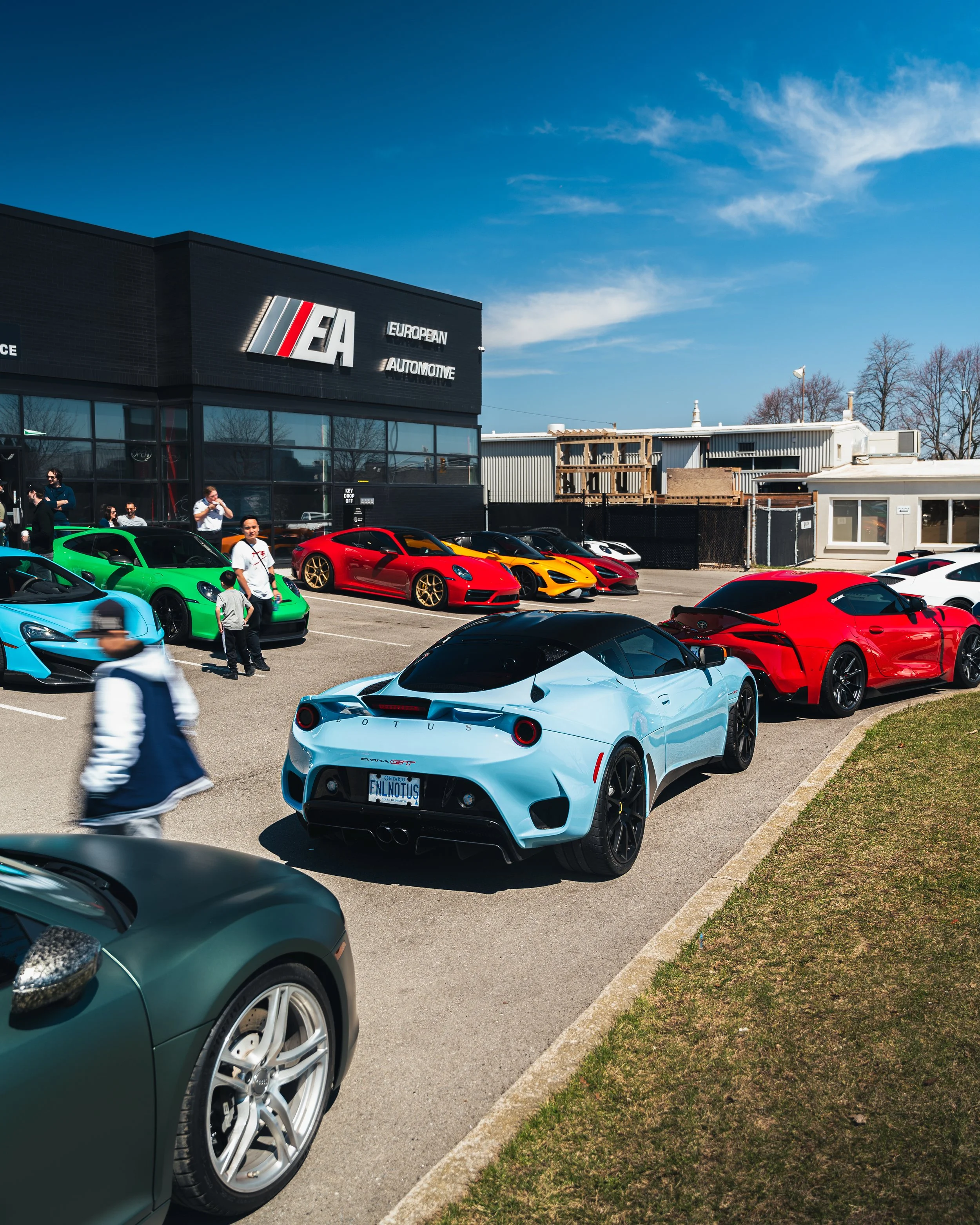 A parking lot filled with colorful high-performance sports cars outside a building with a sign that reads 'European Automotive.' People are walking and socializing around the cars on a clear, sunny day.