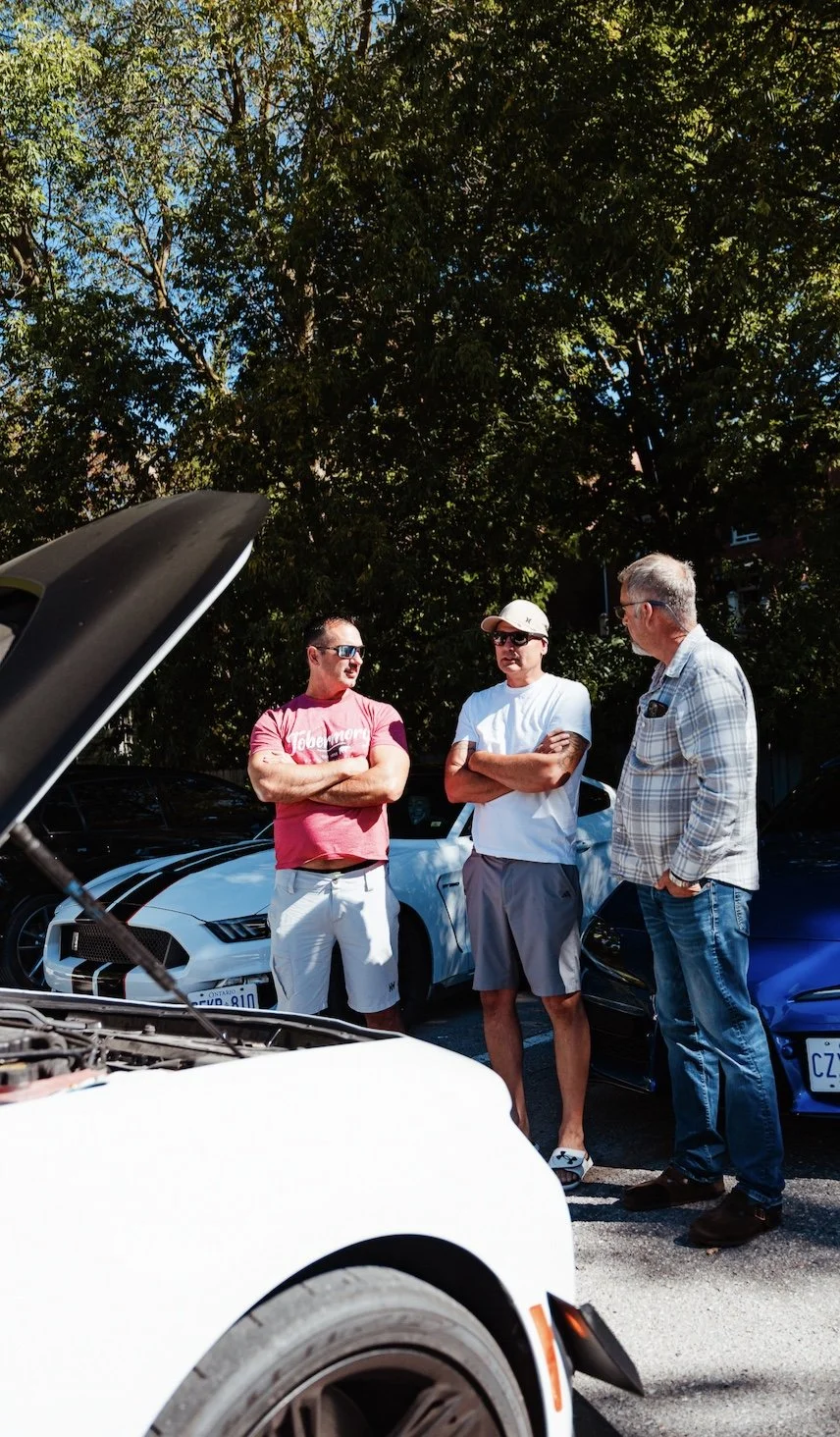 Three men standing next to parked cars outdoors, engaged in conversation, with open car hood visible in the foreground, trees in the background, sunny weather.