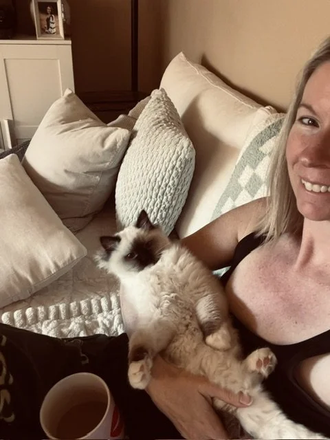 A woman sitting on a bed holding a fluffy Siamese cat with white and brown fur. The bed has multiple pillows and a quilt, with a wooden headboard and framed photos on a white shelf in the background.