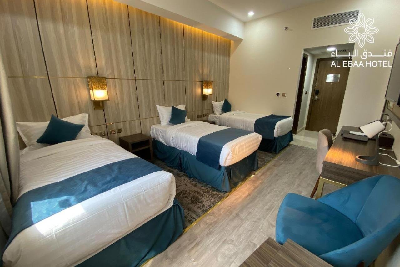 Drei Einzelbetten mit blauen Bettdecken und Kissen in einem Hotelzimmer, Wandlampen an der Wand, Schreibtisch mit Lampe und Stuhl, Teppich auf dem Boden, an der Tür befindet sich ein elektronischer Türschloss. Das Hotel heißt "AL EBAA HOTEL".