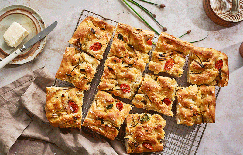 Homemade focaccia bread topped with cherry tomatoes, herbs, and olive oil, cooling on a wire rack.