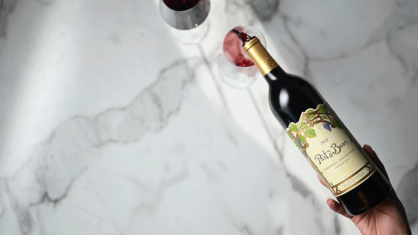 A hand holding a bottle of Post & Beam Cabernet Sauvignon wine with a glass of red wine on a marble surface.