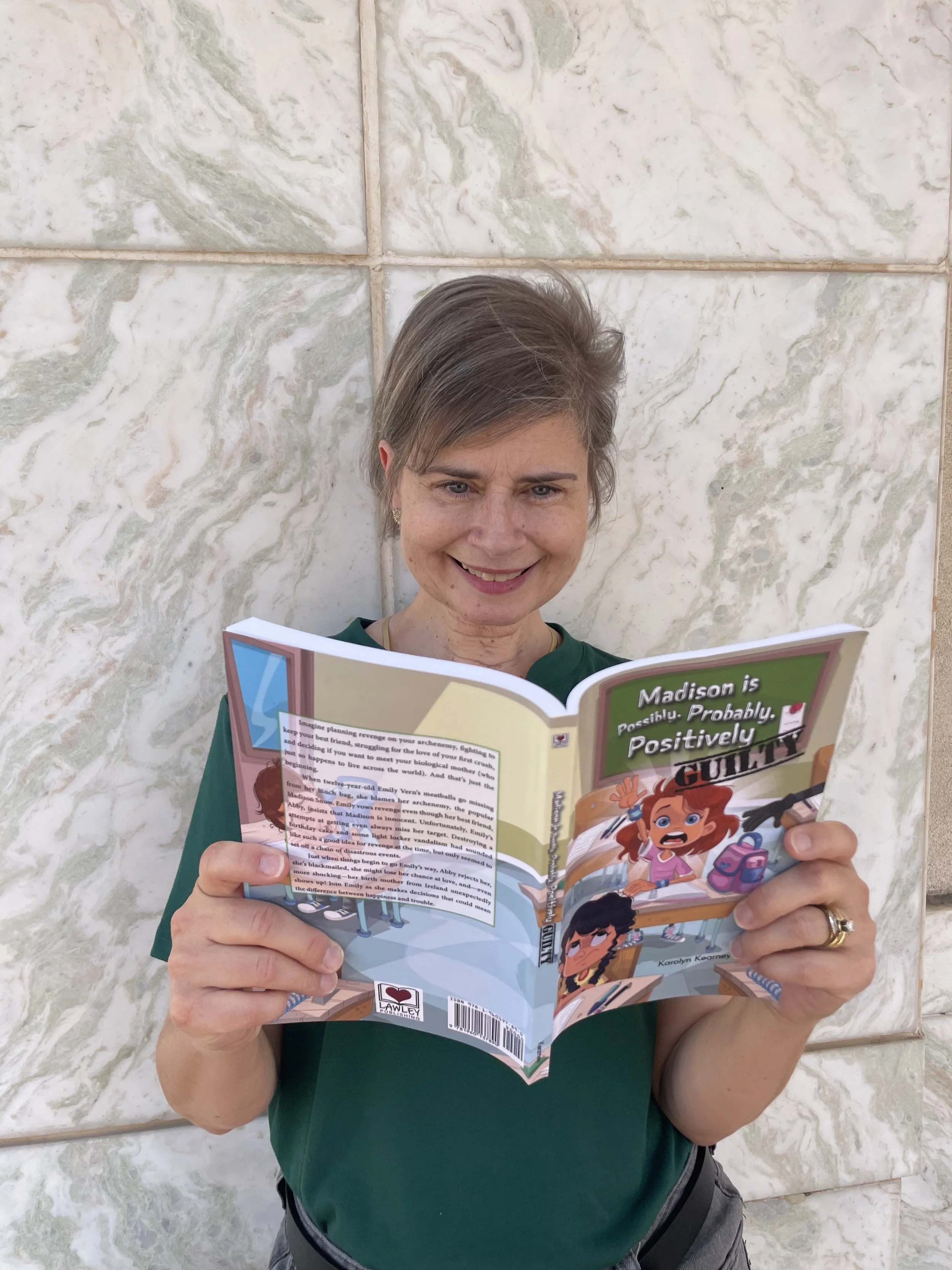 Karolyn Kearney holding and reading her middle grade book Madison Is Possibly, Probably, Positively Guilty against a stone wall background.