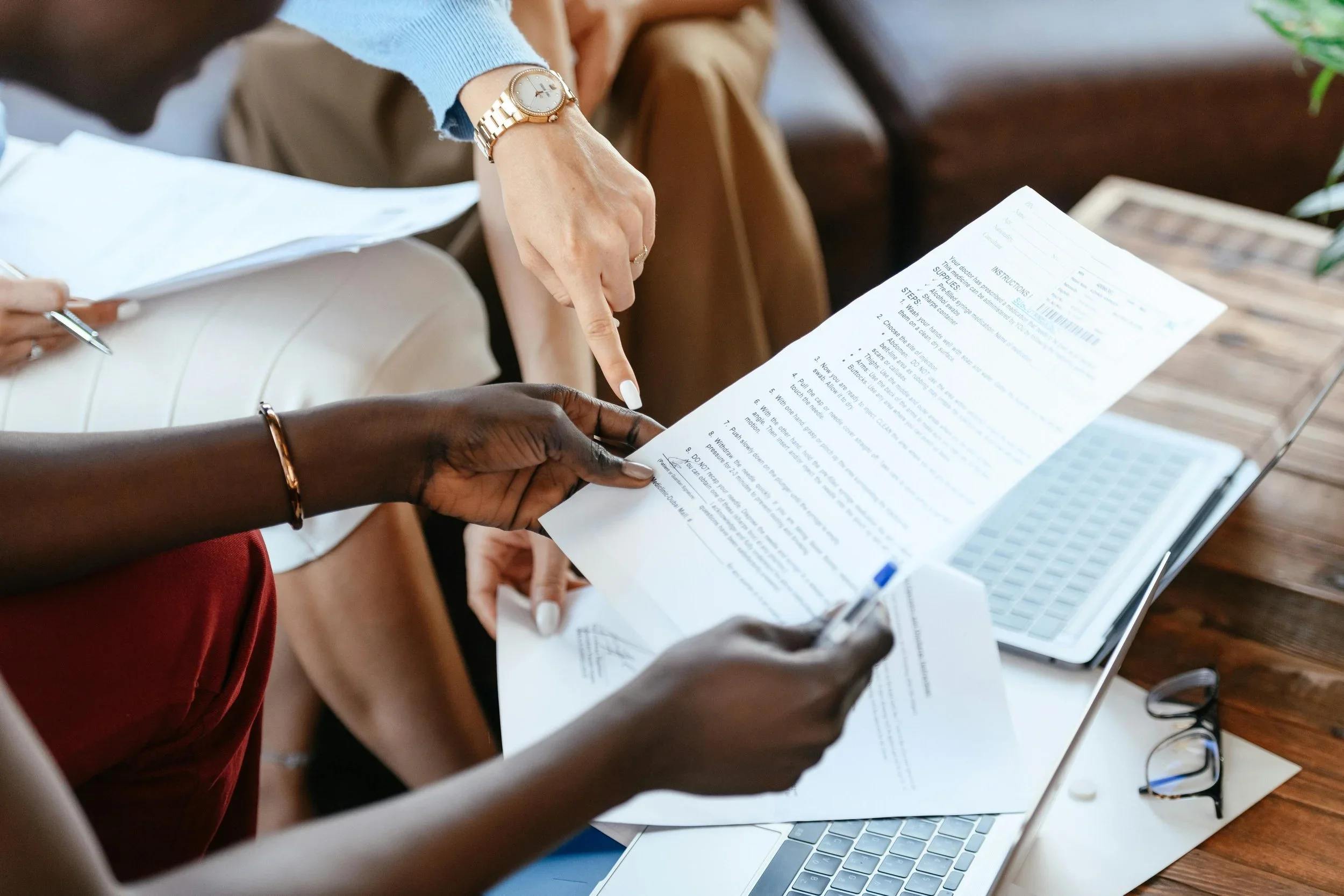 Two people reviewing and discussing paperwork at a wooden table, with a laptop and glasses nearby.