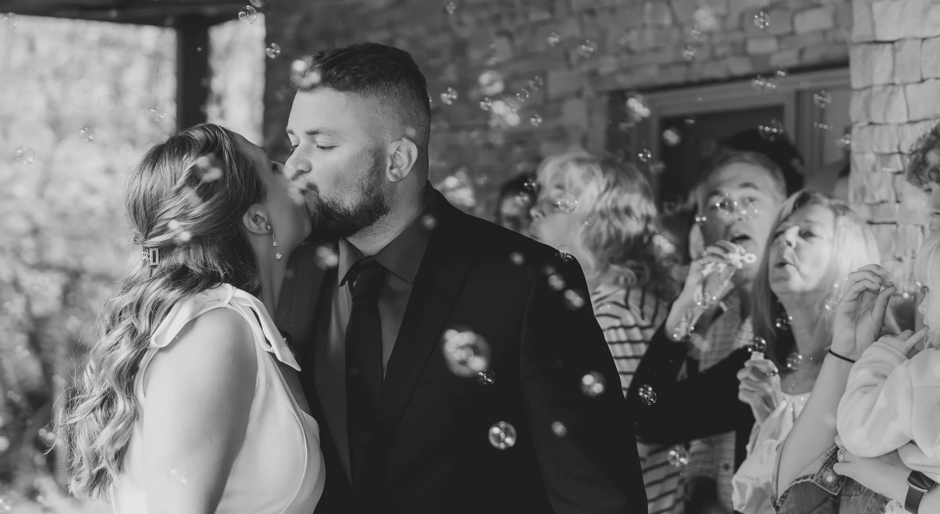 Black and white wedding photo of bride and groom kissing as guests blow bubbles during outdoor ceremony, captured by Maryland wedding photographer