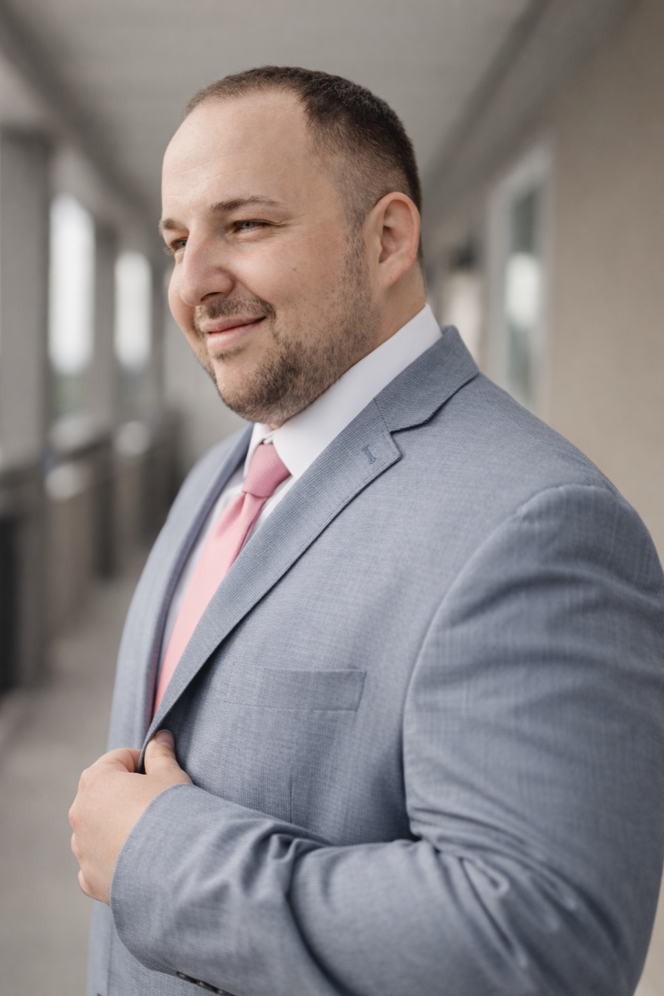 Professional headshot of a confident photographer in a light blue suit with a pink tie, smiling against a soft bokeh background, representing a modern photography business owner.