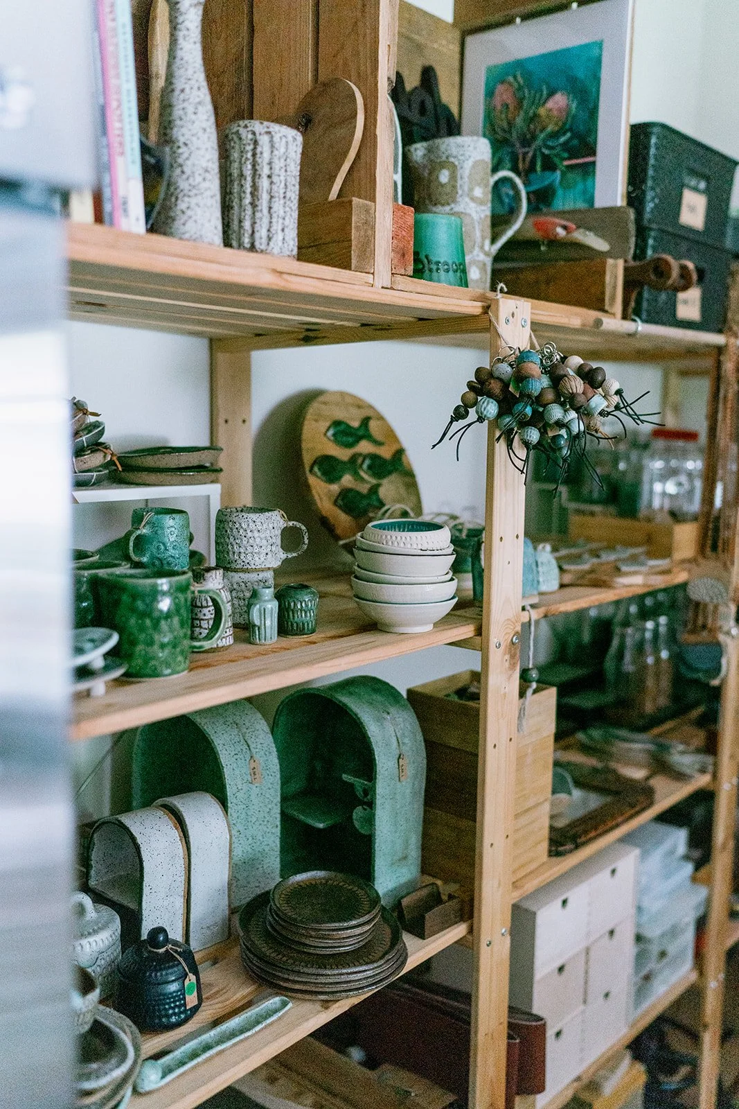Wooden shelving unit filled with pottery, ceramics, and housewares, including plates, bowls, mugs, and decorative items in green, white, and earthy tones.