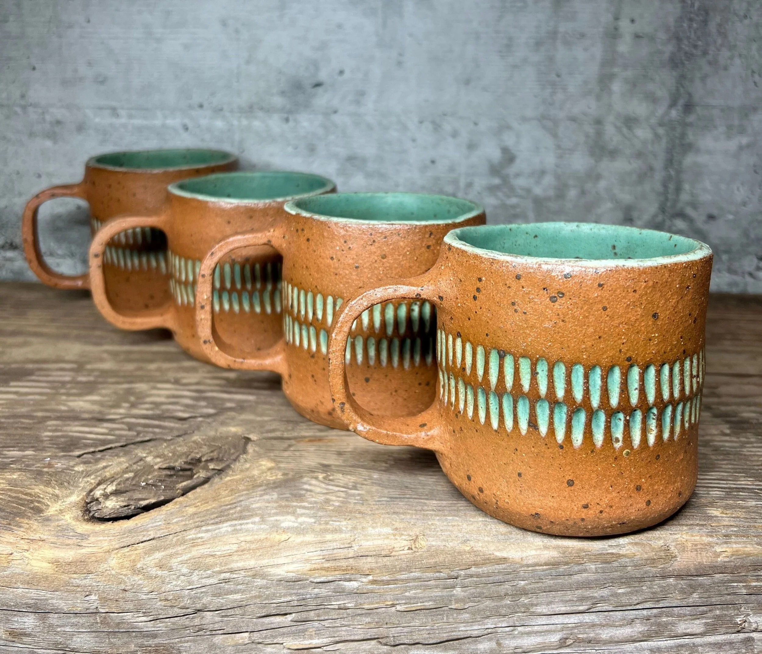 Four matching ceramic coffee mugs with a textured brown exterior and turquoise interior, arranged in a diagonal line on a rustic wooden surface with a gray concrete wall background.