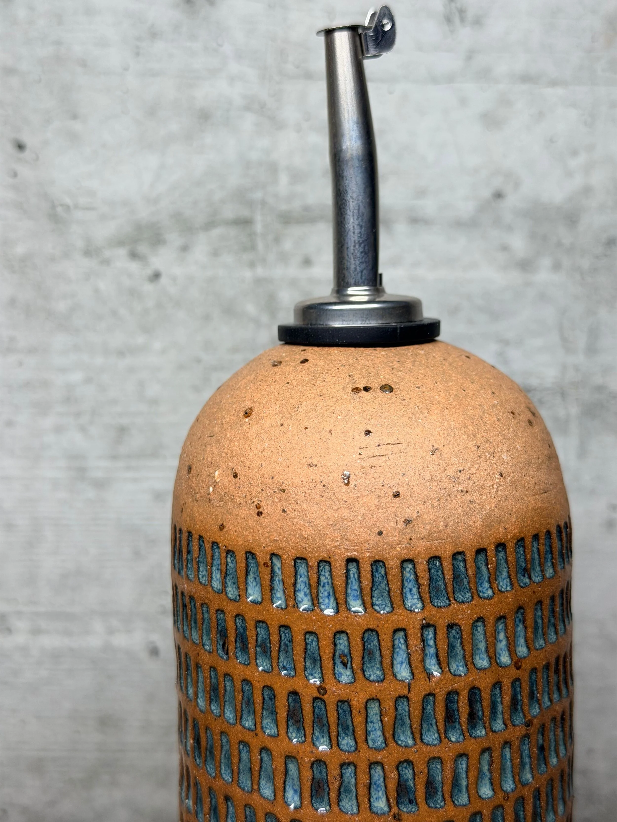 Handmade from stoneware clay, it features a textured pattern and is fitted with a stainless steel pourer for smooth, controlled dispensing.