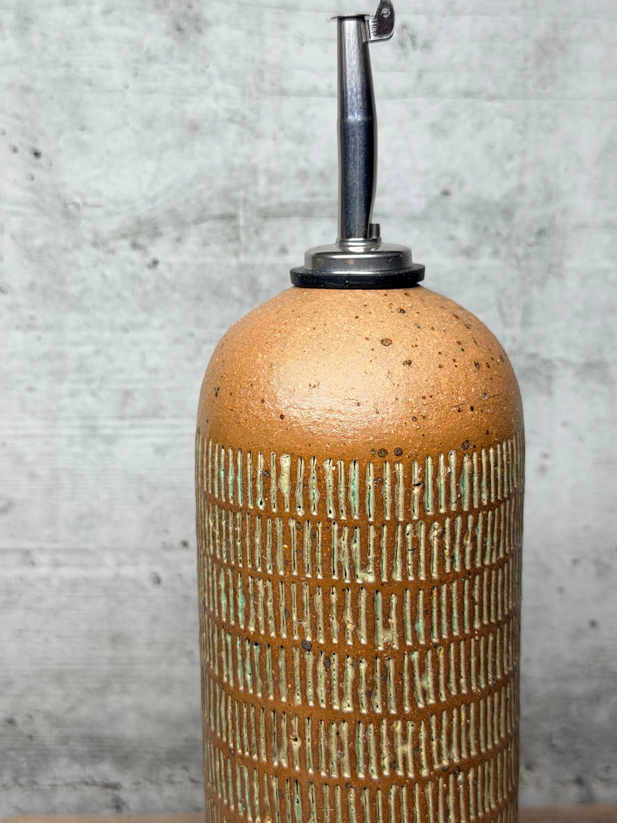 Handmade from stoneware clay, it features a textured pattern and is fitted with a stainless steel pourer for smooth, controlled dispensing.