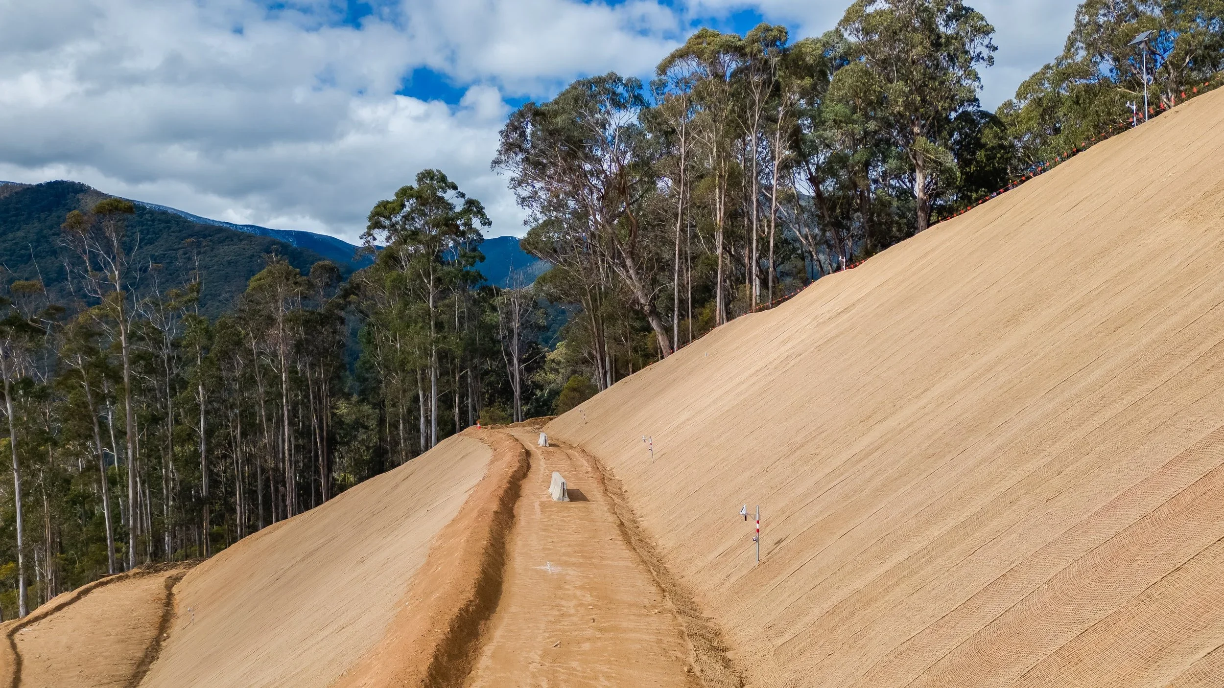 Bogong High Plains landslip construction site with a dirt path winding up a large, sloped area covered in soil, with trees in the background and a partly cloudy sky.