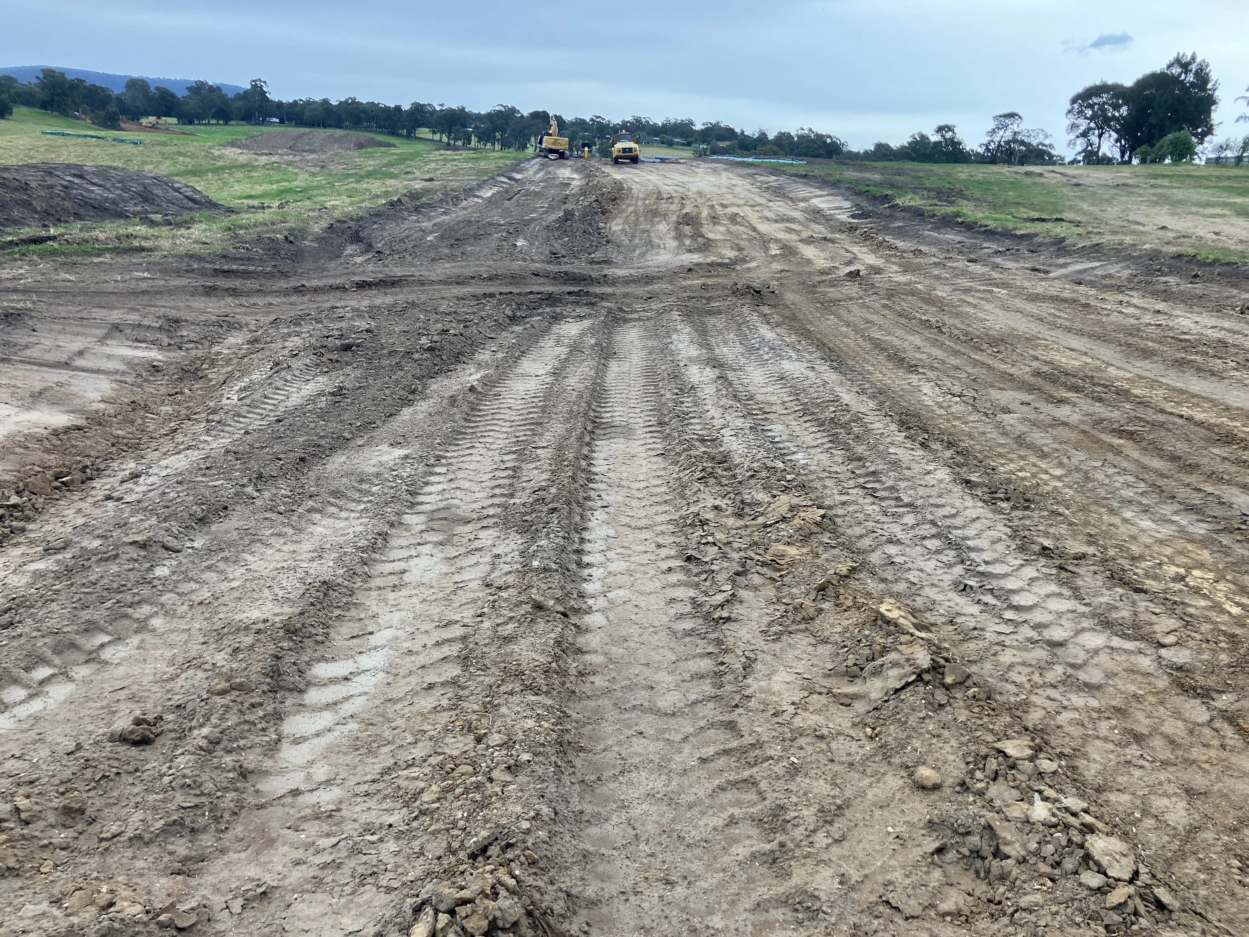 A dirt road under construction with construction equipment in the background and scattered green grassy areas on either side.