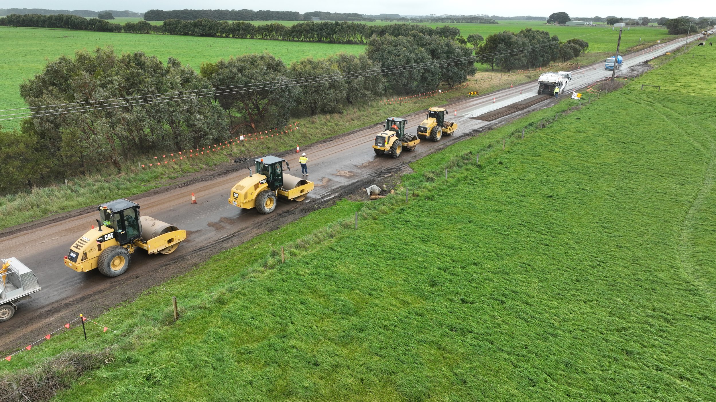 Construction workers and heavy machinery are paving a rural road across green fields with trees in the background.