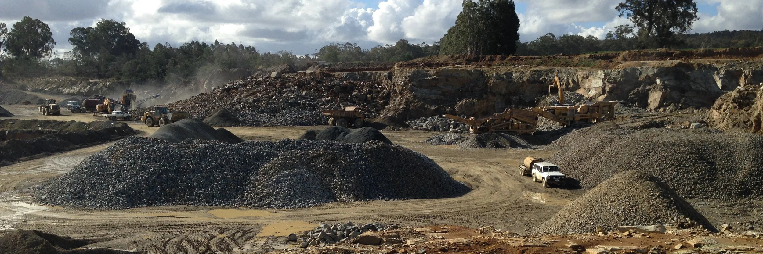 A construction site or quarry with multiple large quantities of gravel, rocks, and dirt, with heavy machinery and trucks working on excavation and transportation, set against a partly cloudy sky and forested hillside.