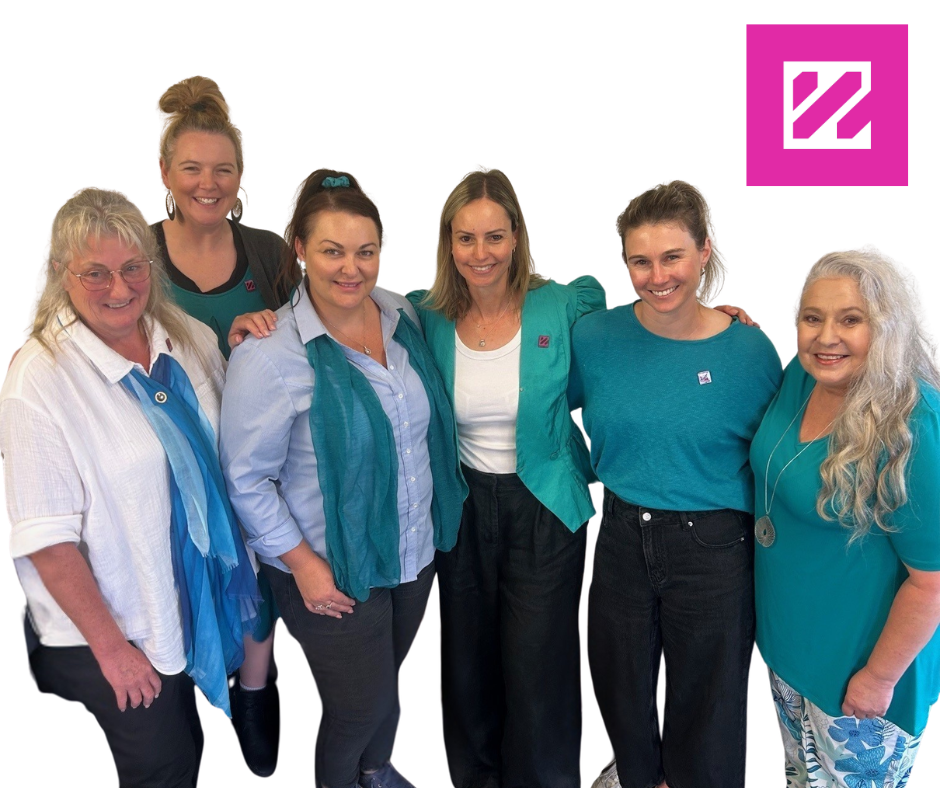 Group of six women smiling with arm around each other, wearing teal and blue clothing, standing against a white background with a pink WOW logo in the top right corner.