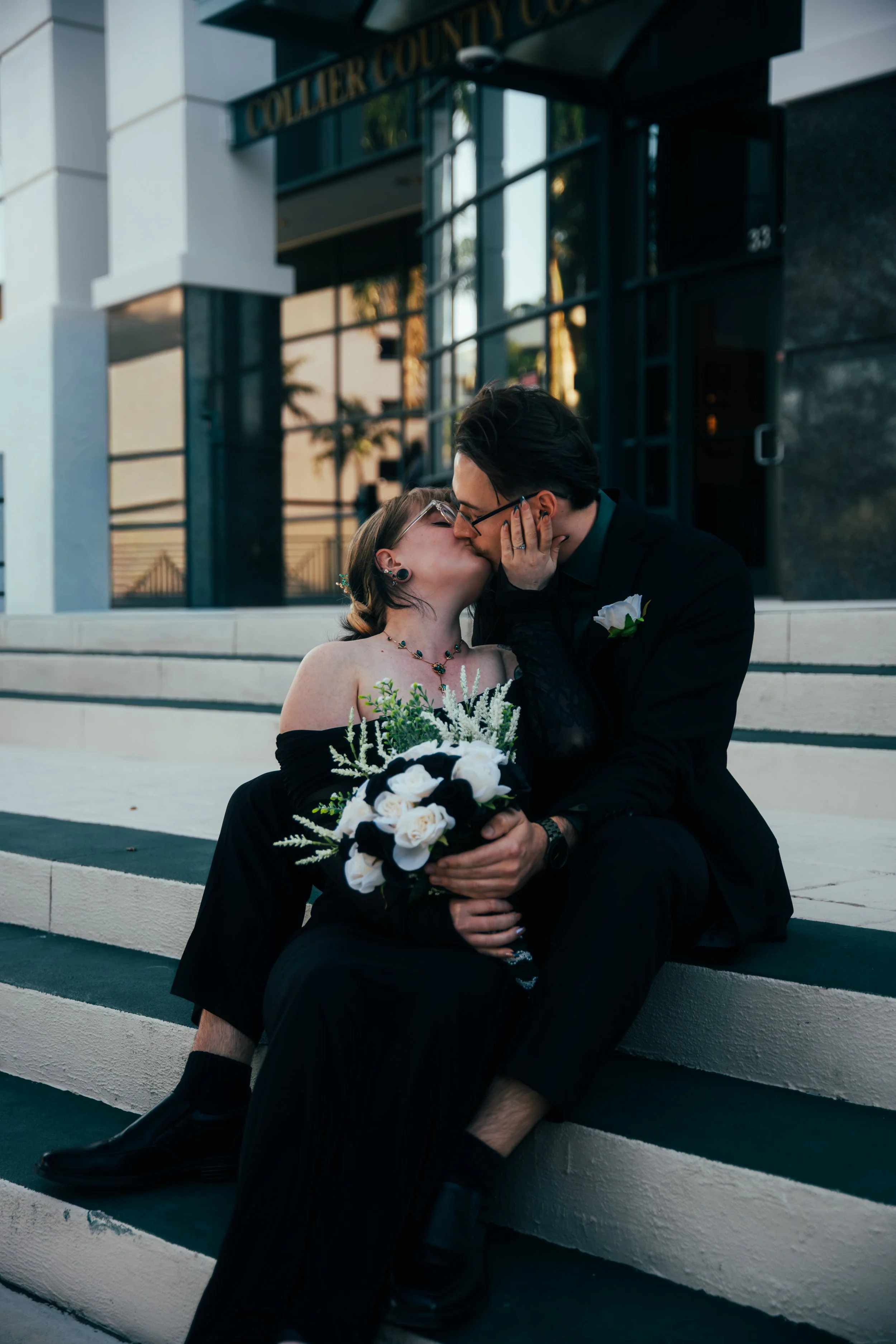 A couple sharing a kiss on the steps outside a wedding venue, with the woman holding a bouquet of white and black flowers. After wedding photos