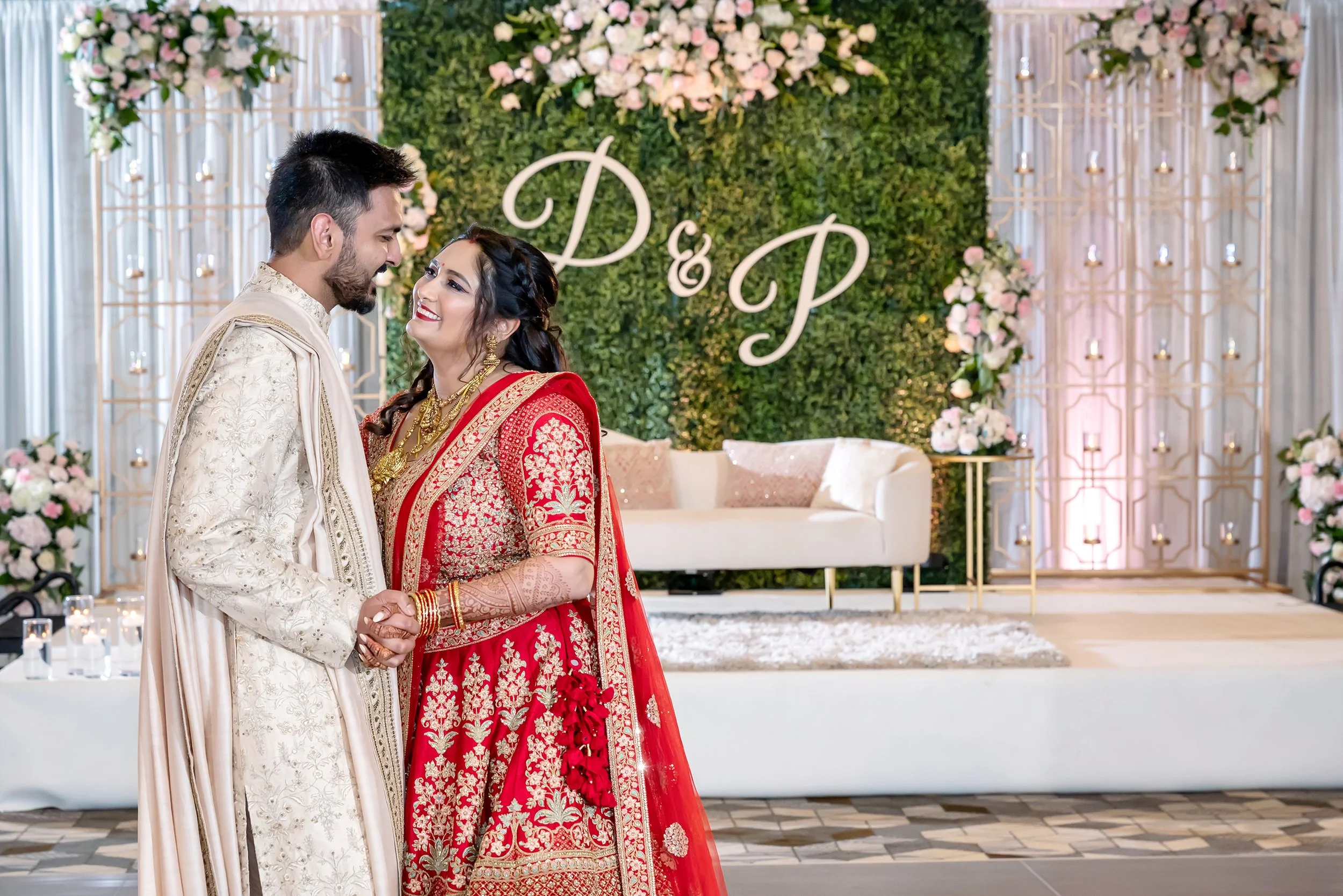 Bride and groom in traditional attire at a wedding venue with floral decor and initials "D & P"