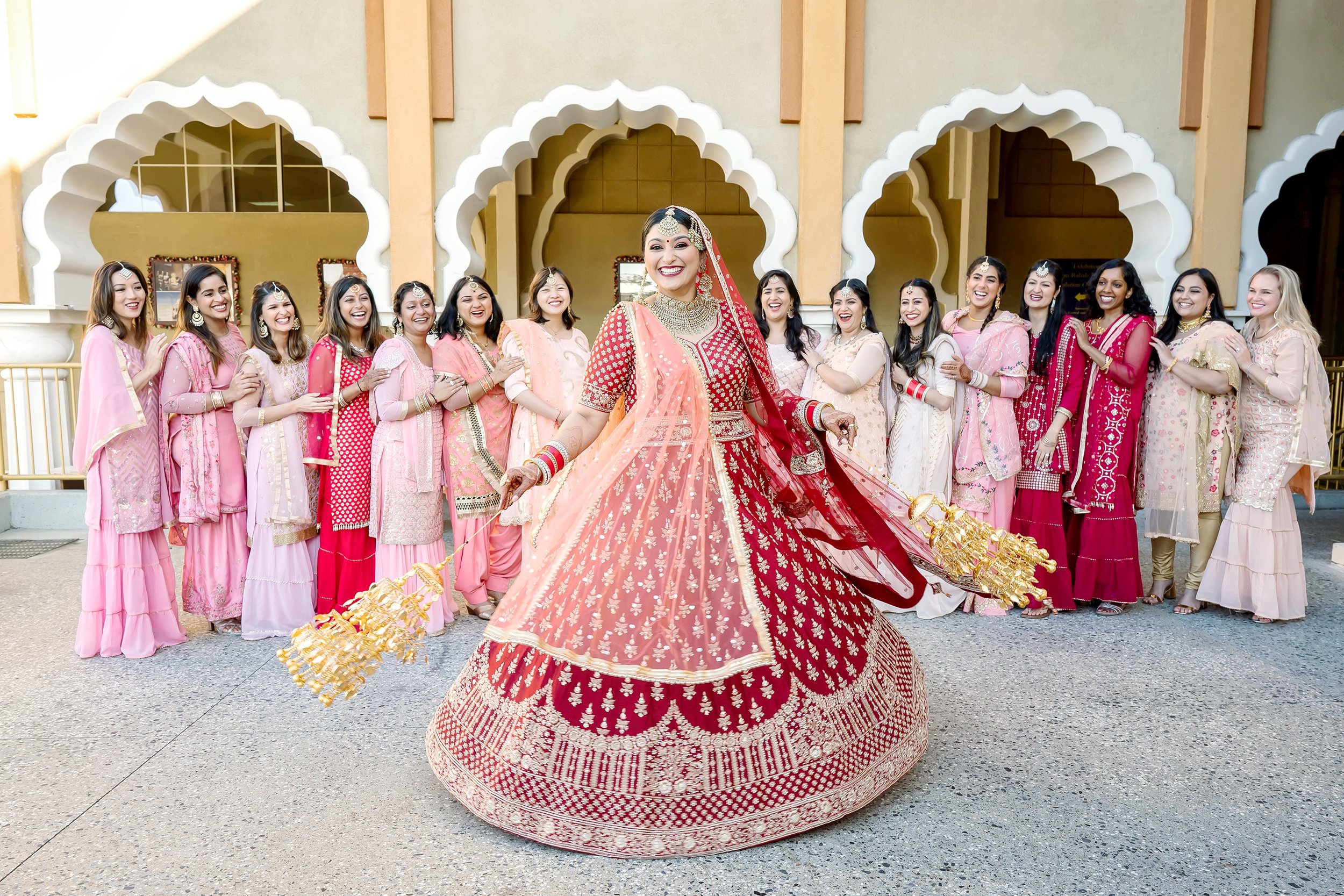 Bride in red traditional Indian bridal attire, surrounded by bridesmaids in pink outfits, posing happily in front of an ornate archway backdrop.