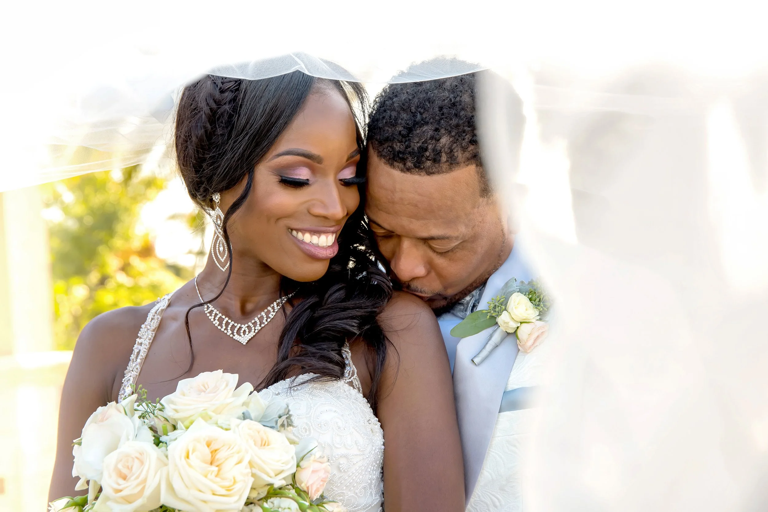 Bride and groom embracing under veil, smiling, holding bouquet of white roses.