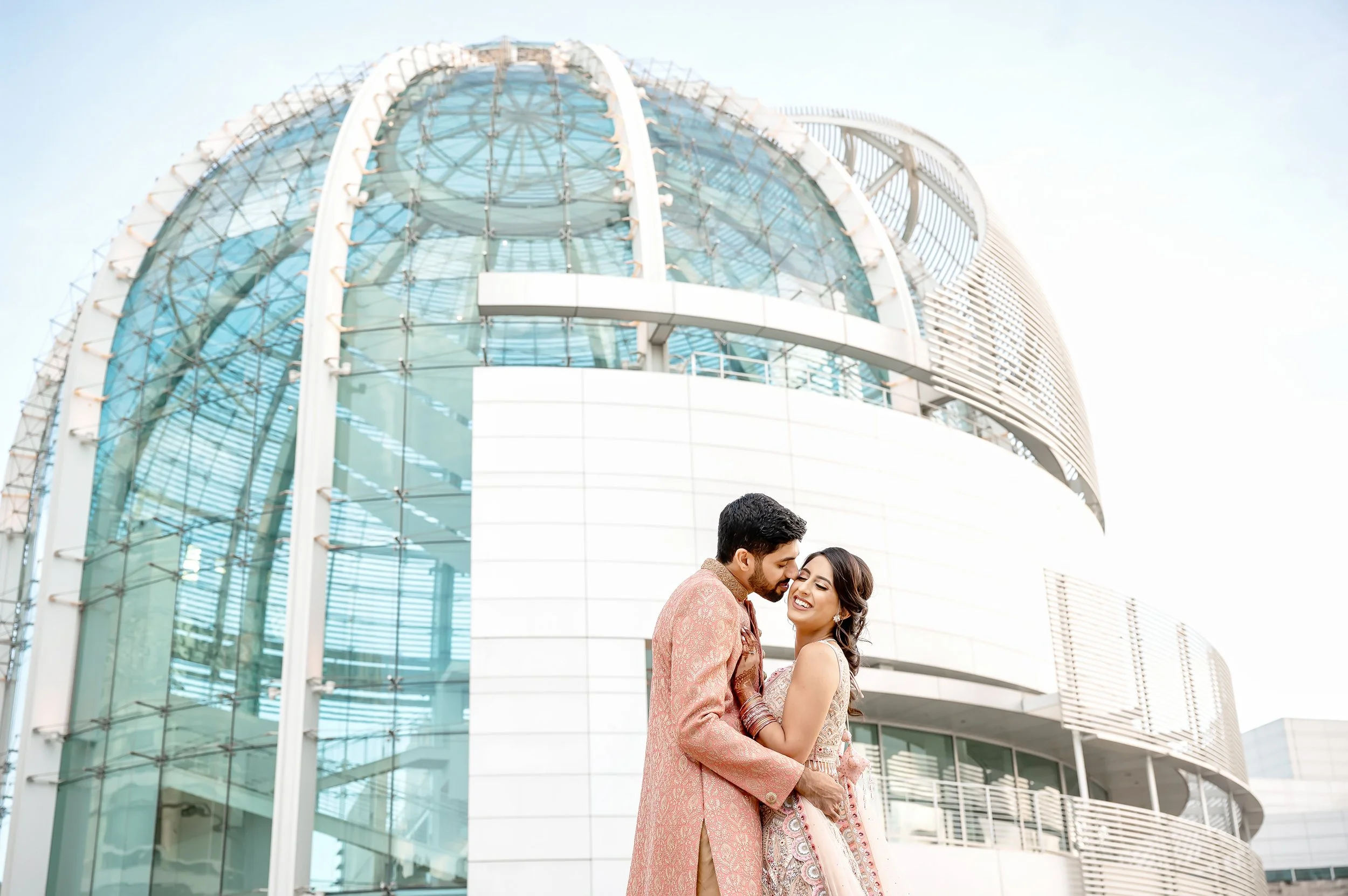Couple in traditional attire embracing in front of a modern glass-domed building.