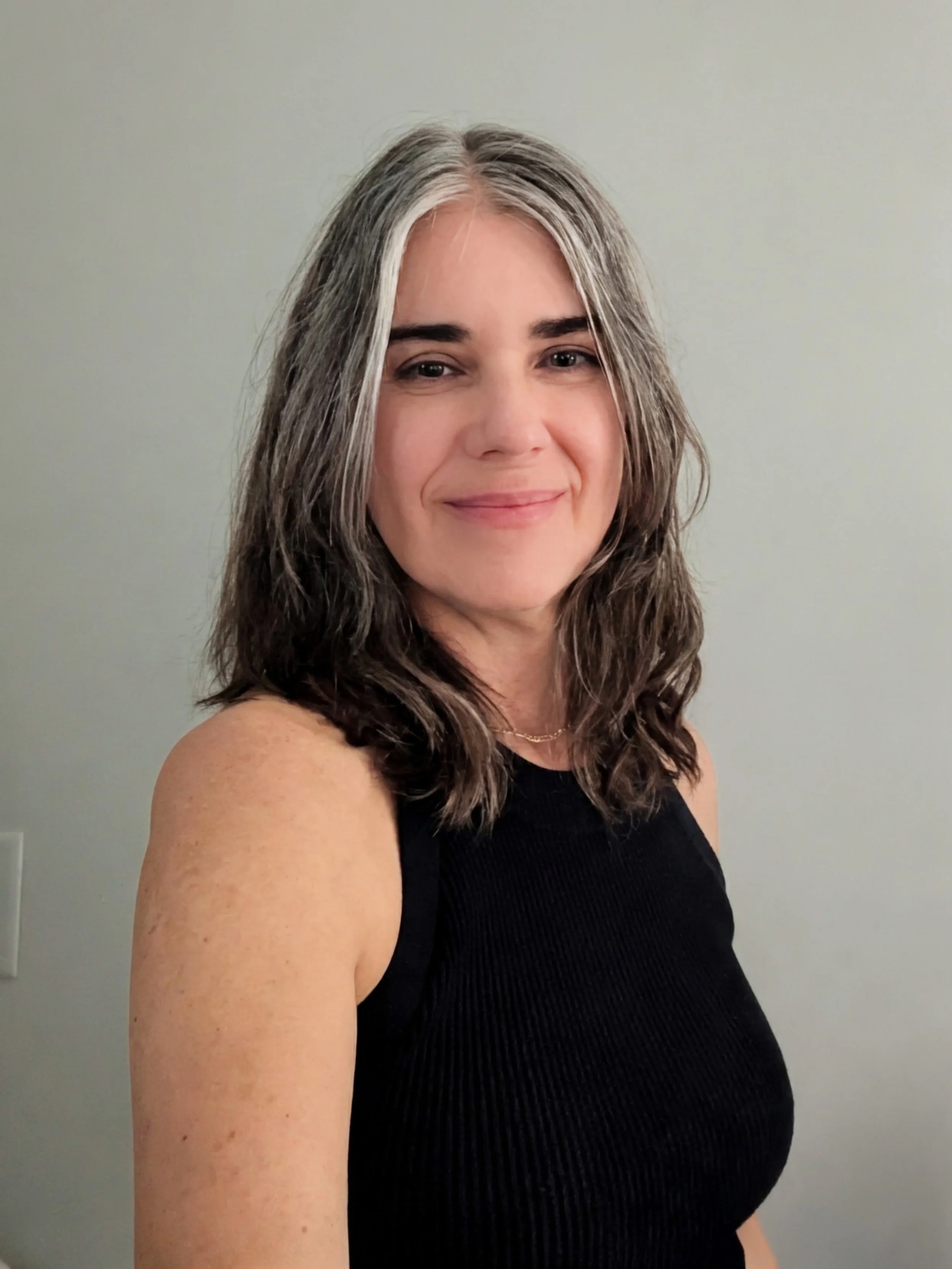A smiling woman with shoulder-length hair with gray streaks, wearing a black sleeveless top, standing against a plain light-colored wall.
