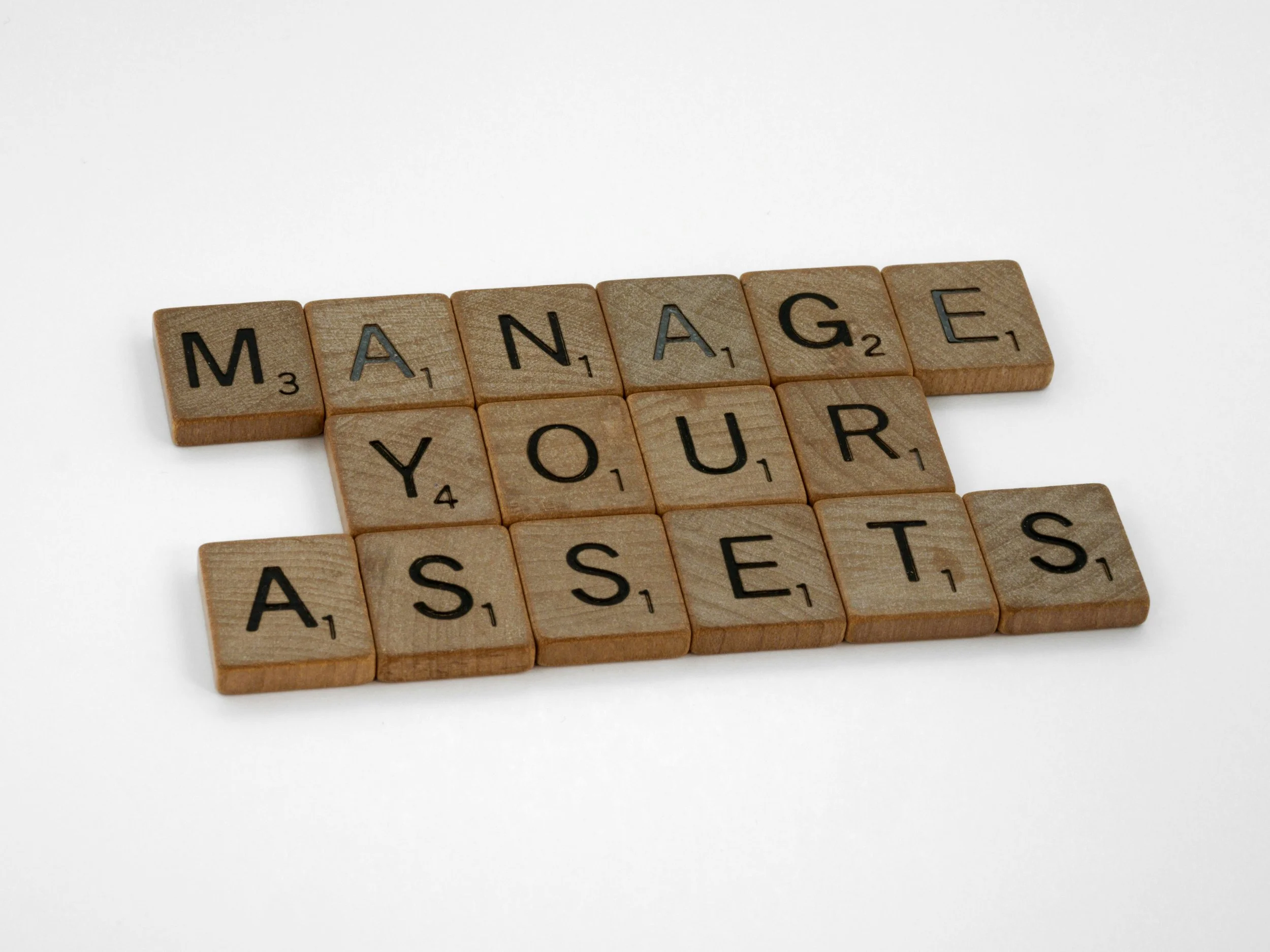 Wooden Scrabble tiles spelling out "MANAGE YOUR ASSETS" on a white background.
