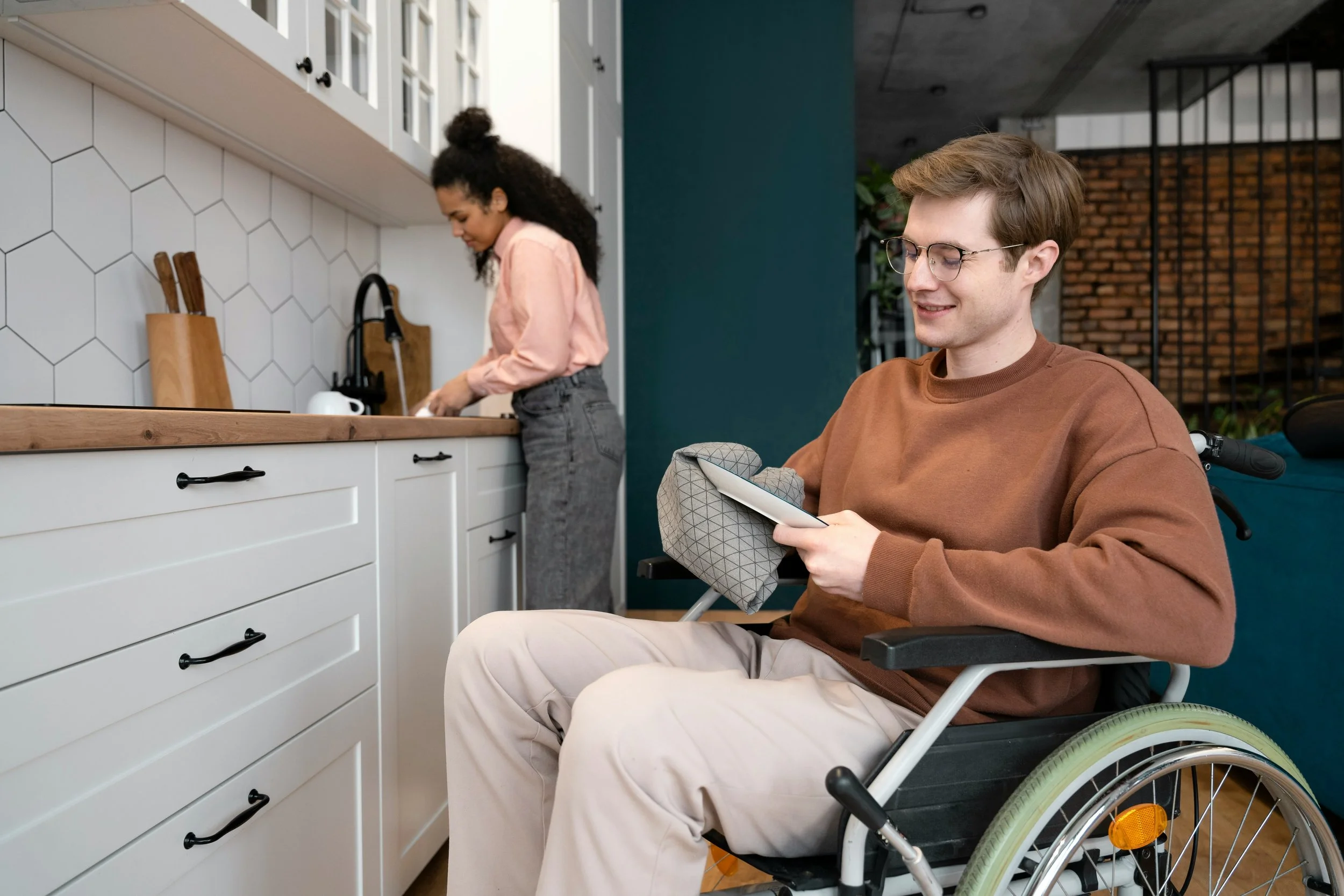 A woman with curly hair wearing a pink shirt and gray jeans is washing dishes at the kitchen sink. A man with glasses and brown hair, wearing a brown sweatshirt and beige pants, is sitting in a wheelchair, holding a towel and a tablet, smiling in the foreground.