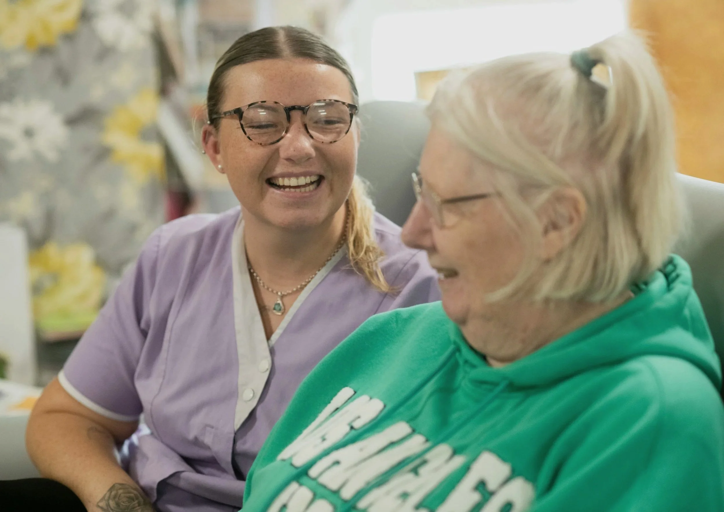 A young woman with glasses and a tattoo on her left arm smiling and talking to an elderly woman with glasses and a ponytail, both in a healthcare setting.