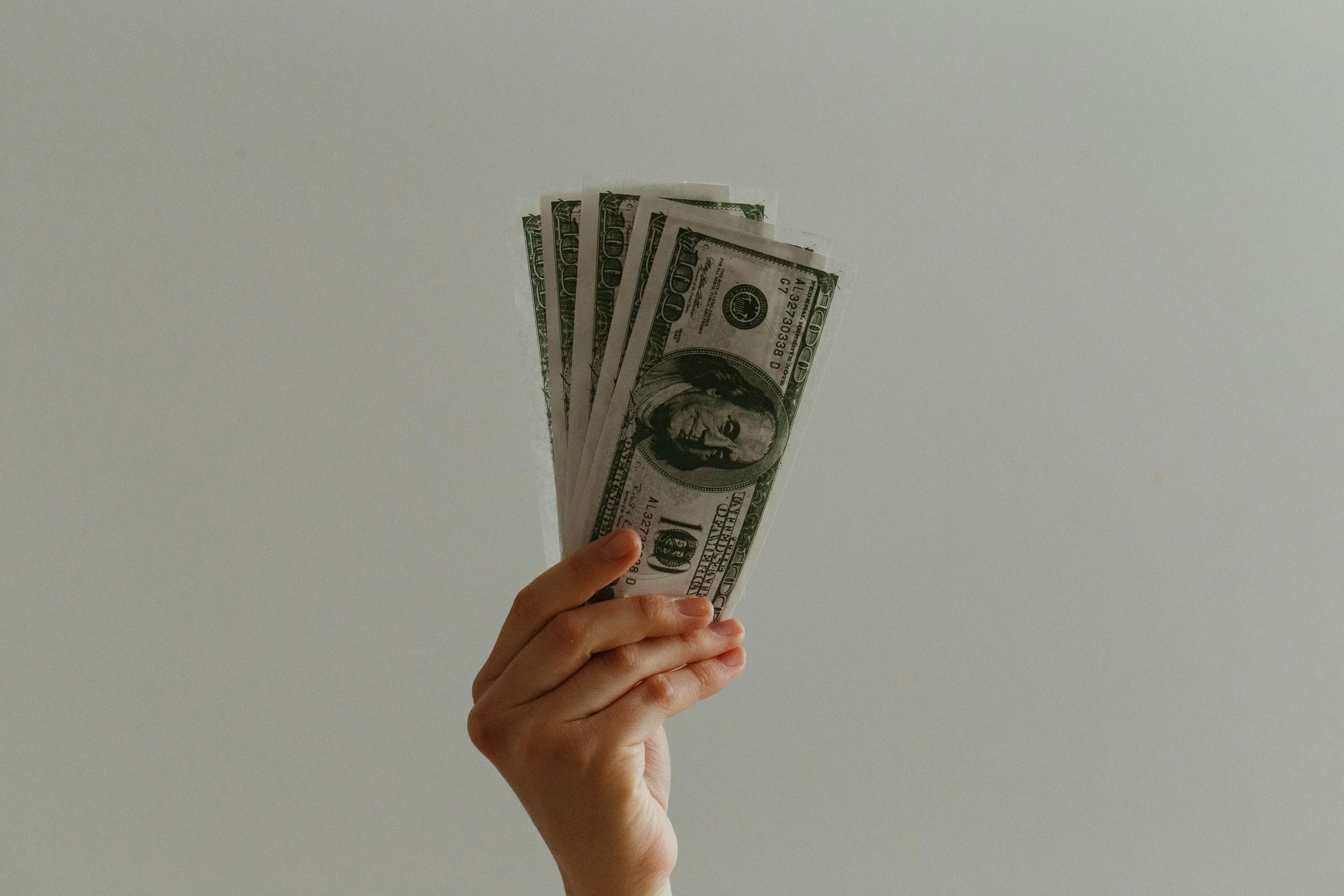 A hand holding a fanned stack of five US hundred-dollar bills against a plain white background.